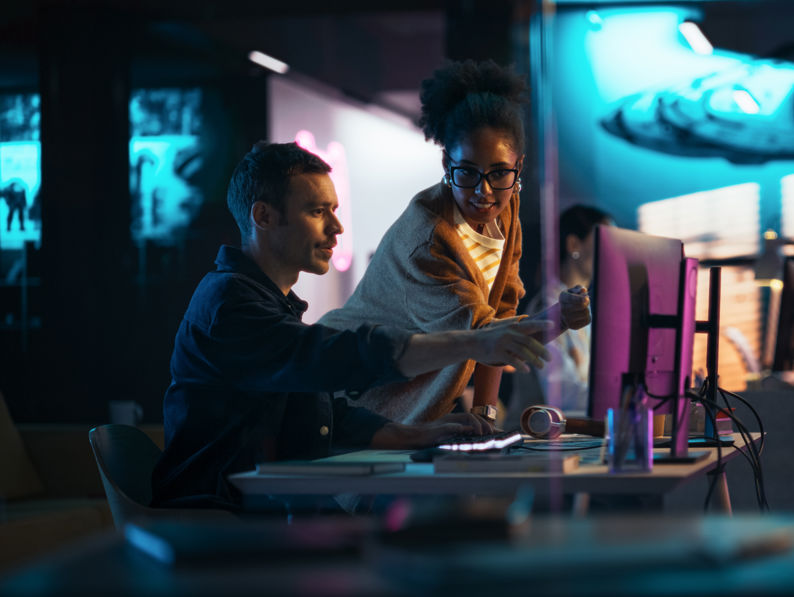 Two people collaborate at a computer in a dimly lit, modern office with sci-fi spaceship artwork on the wall. One person points at the monitor while the other stands, both focused on the screen.