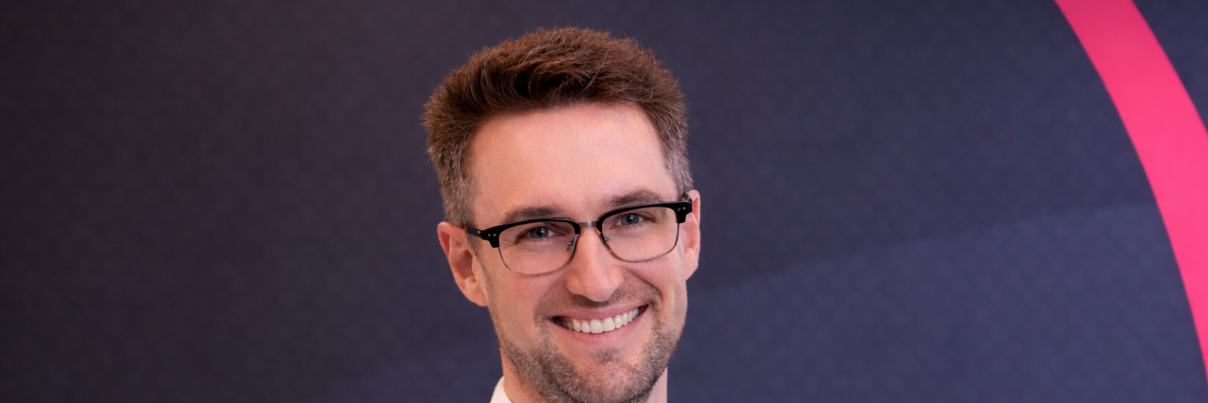 A man with short brown hair and glasses, wearing a white button-up shirt, smiles at the camera. The background is dark with geometric patterns and a partial pink arc.