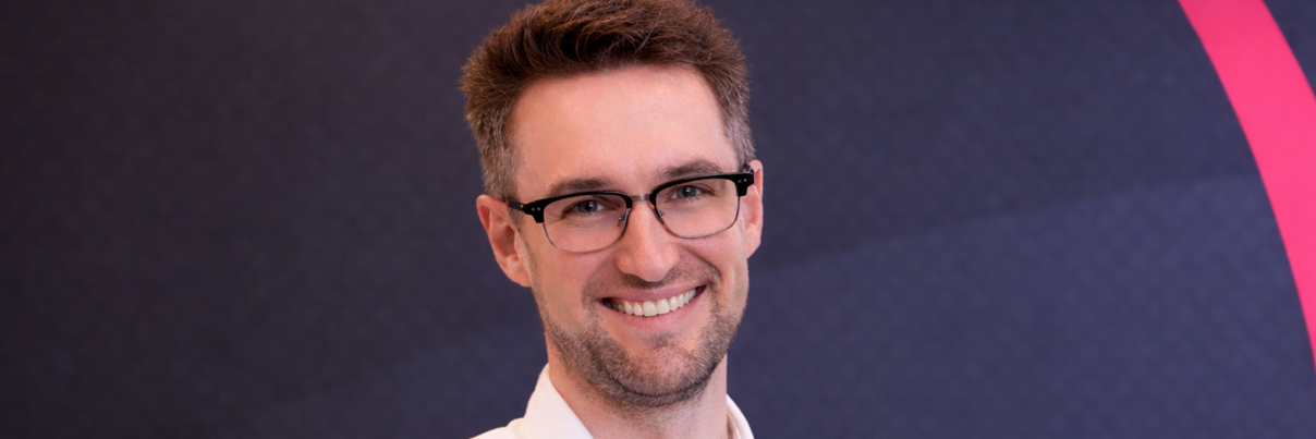 A man with short brown hair and glasses, wearing a white button-up shirt, smiles at the camera. The background is dark with geometric patterns and a partial pink arc.