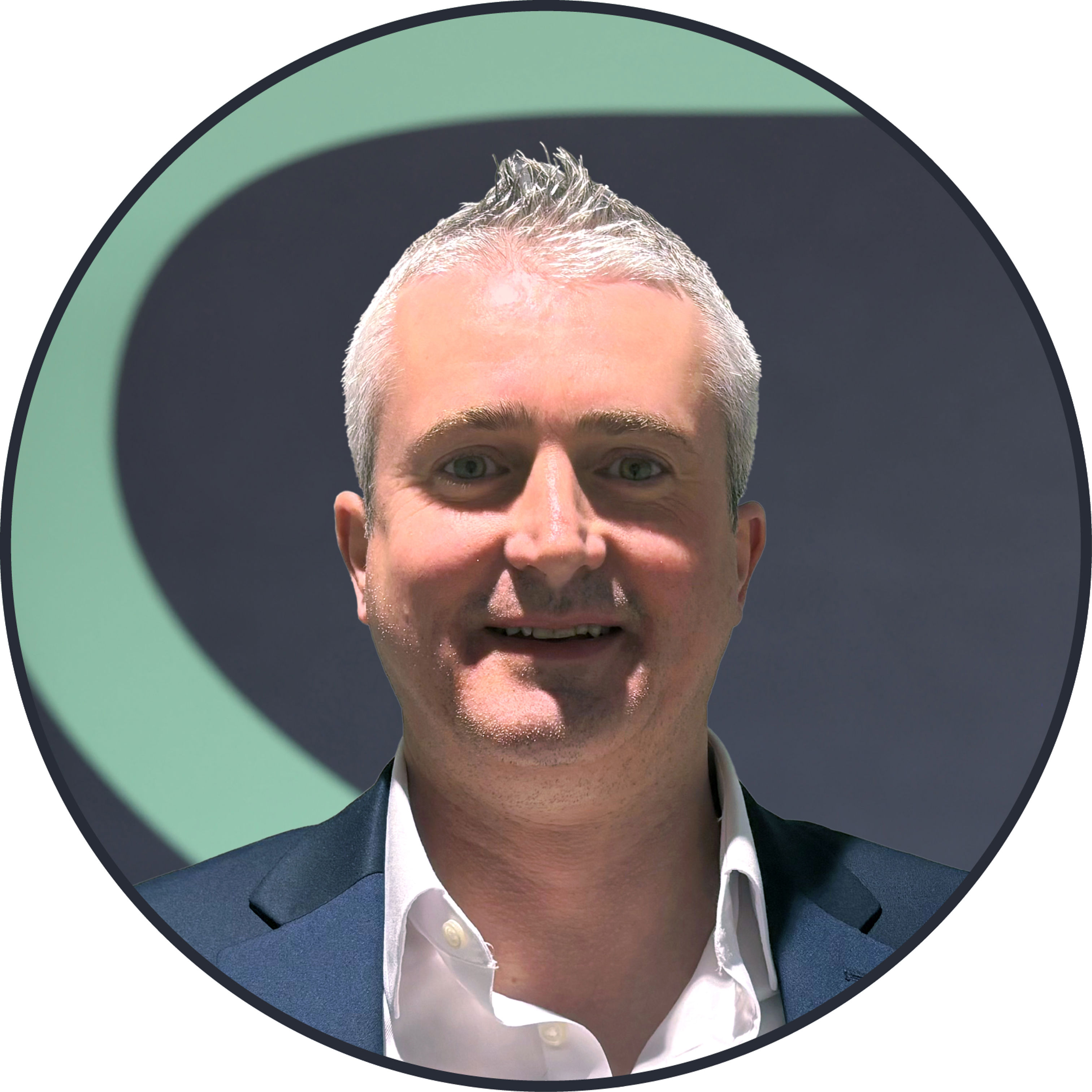 A man with short gray hair wearing a dark suit and white shirt, smiling, is shown against a dark background with a large green curved shape. The image is framed in a circular border.