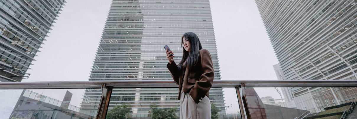 A woman stands on an outdoor terrace, looking at her phone, with three tall modern glass buildings towering above her against a light sky.