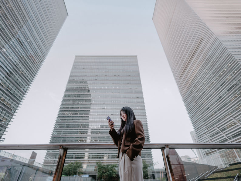 A woman stands on an outdoor terrace, looking at her phone, with three tall modern glass buildings towering above her against a light sky.