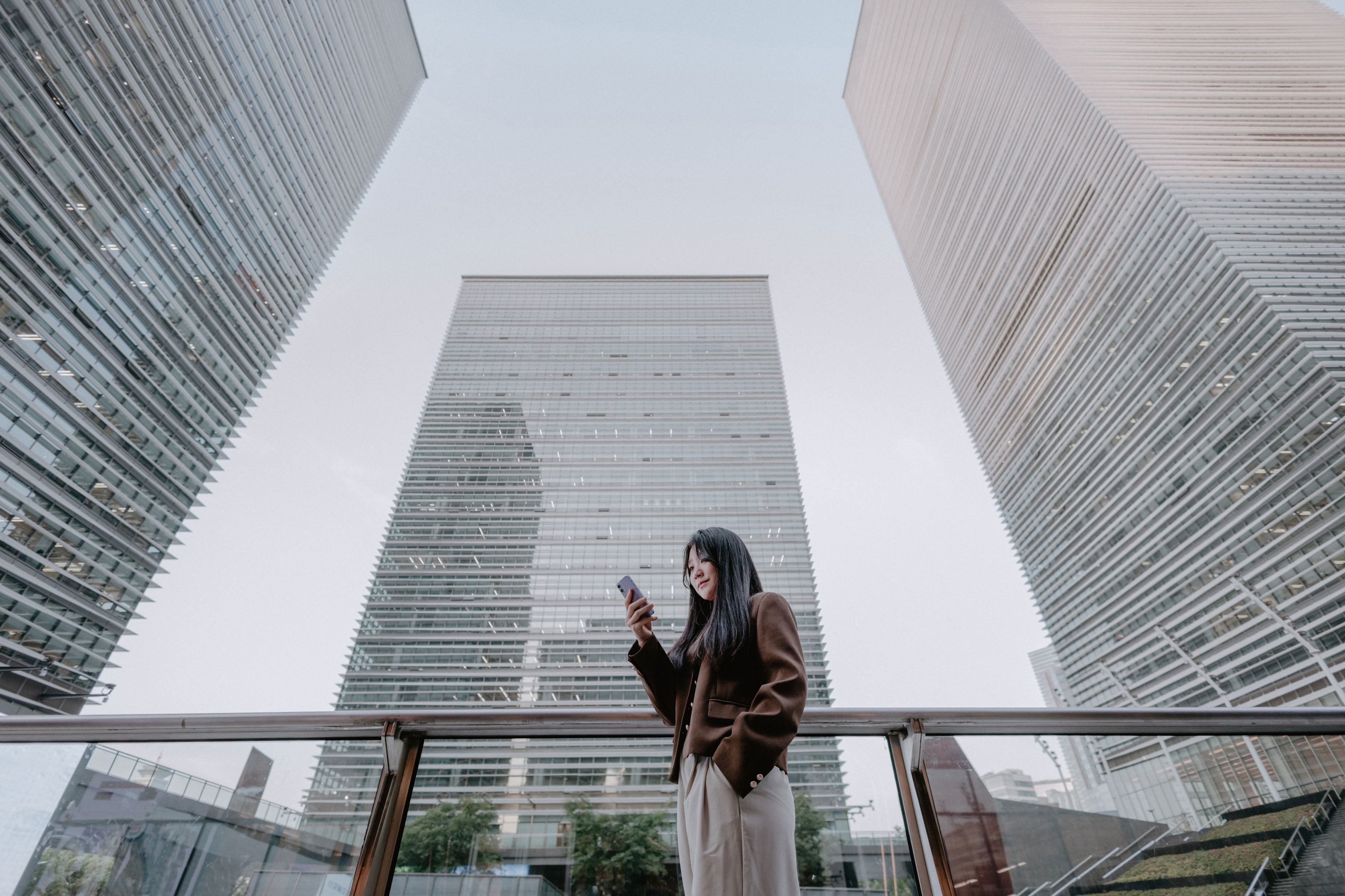 A woman stands on an outdoor terrace, looking at her phone, with three tall modern glass buildings towering above her against a light sky.