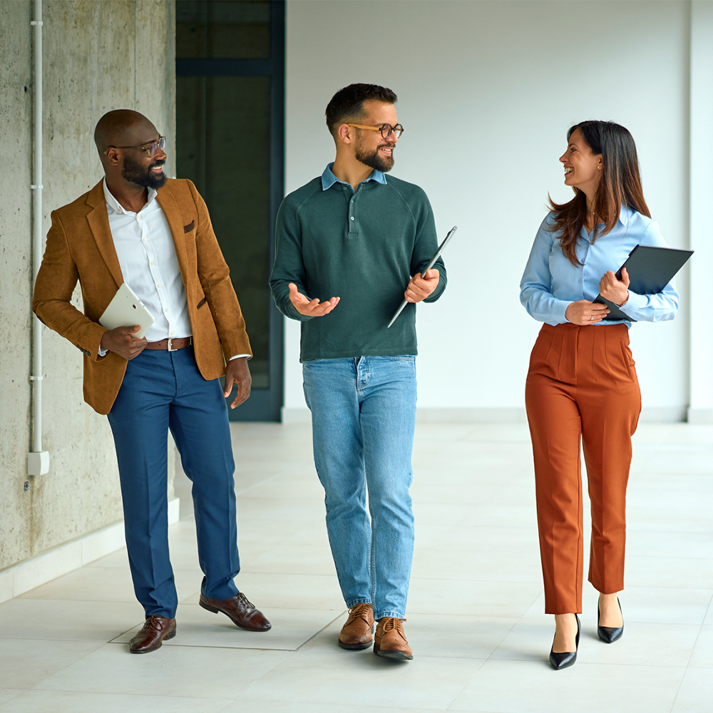 Three colleagues in business-casual attire walk through an office building while talking and holding folders and a tablet.