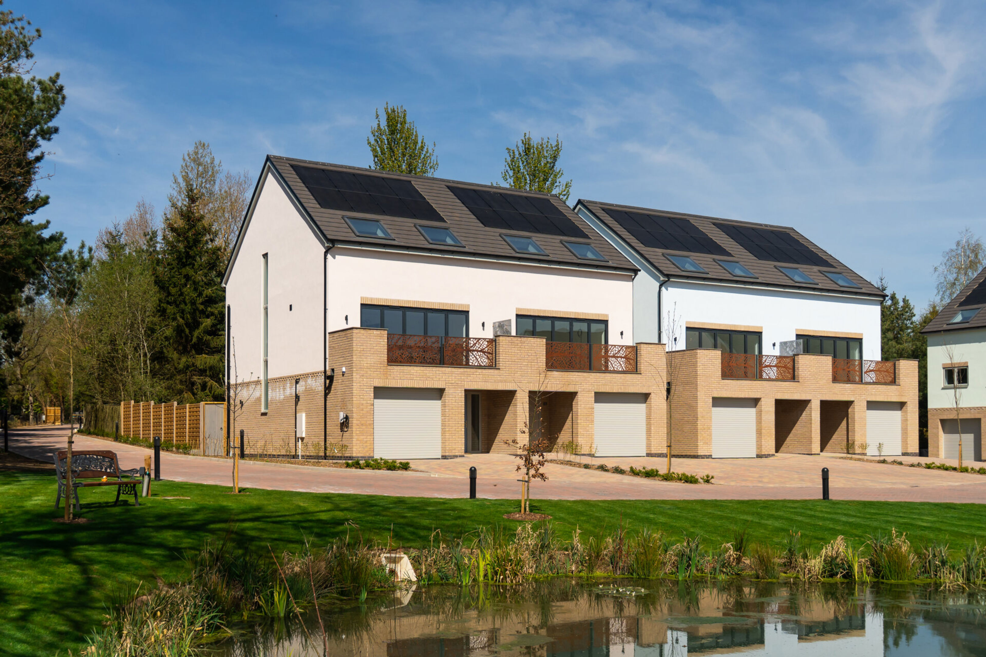 Modern row of three-story townhouses with solar panels on the roofs, large windows, and garages. There is a pond and green lawn in the foreground, with trees and blue sky in the background.