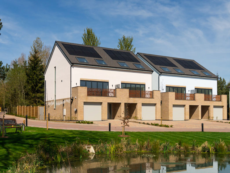 Modern row of three-story townhouses with solar panels on the roofs, large windows, and garages. There is a pond and green lawn in the foreground, with trees and blue sky in the background.
