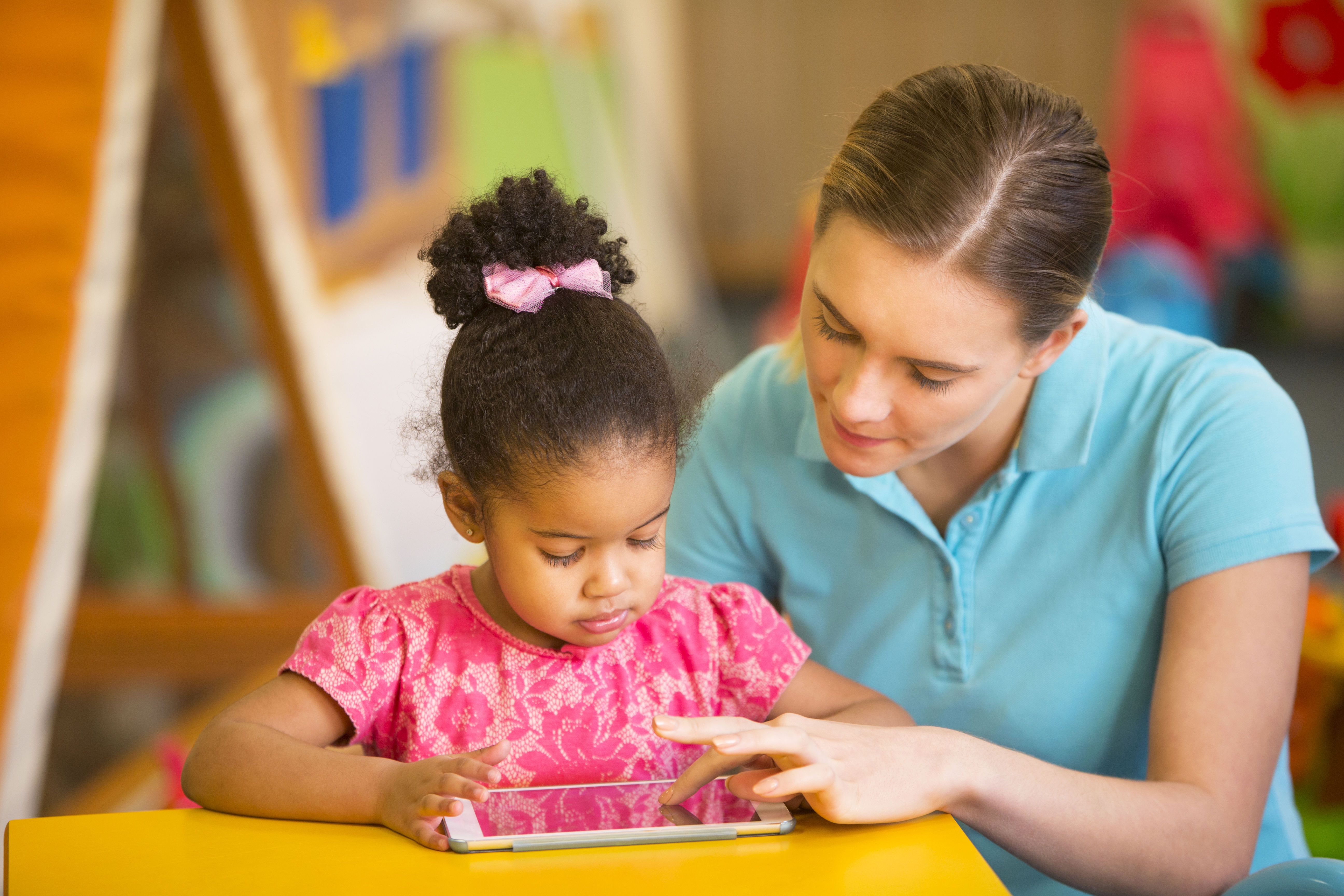 A woman helps a young girl use a tablet at a colorful table in a classroom or playroom setting.