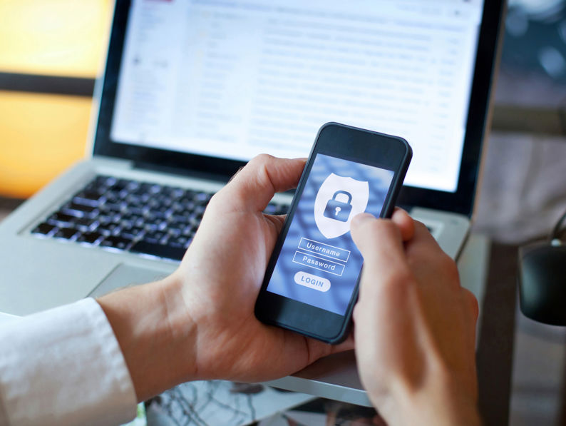 A person holds a smartphone displaying a login screen with username and password fields, in front of an open laptop showing an email inbox, suggesting digital security or online authentication.