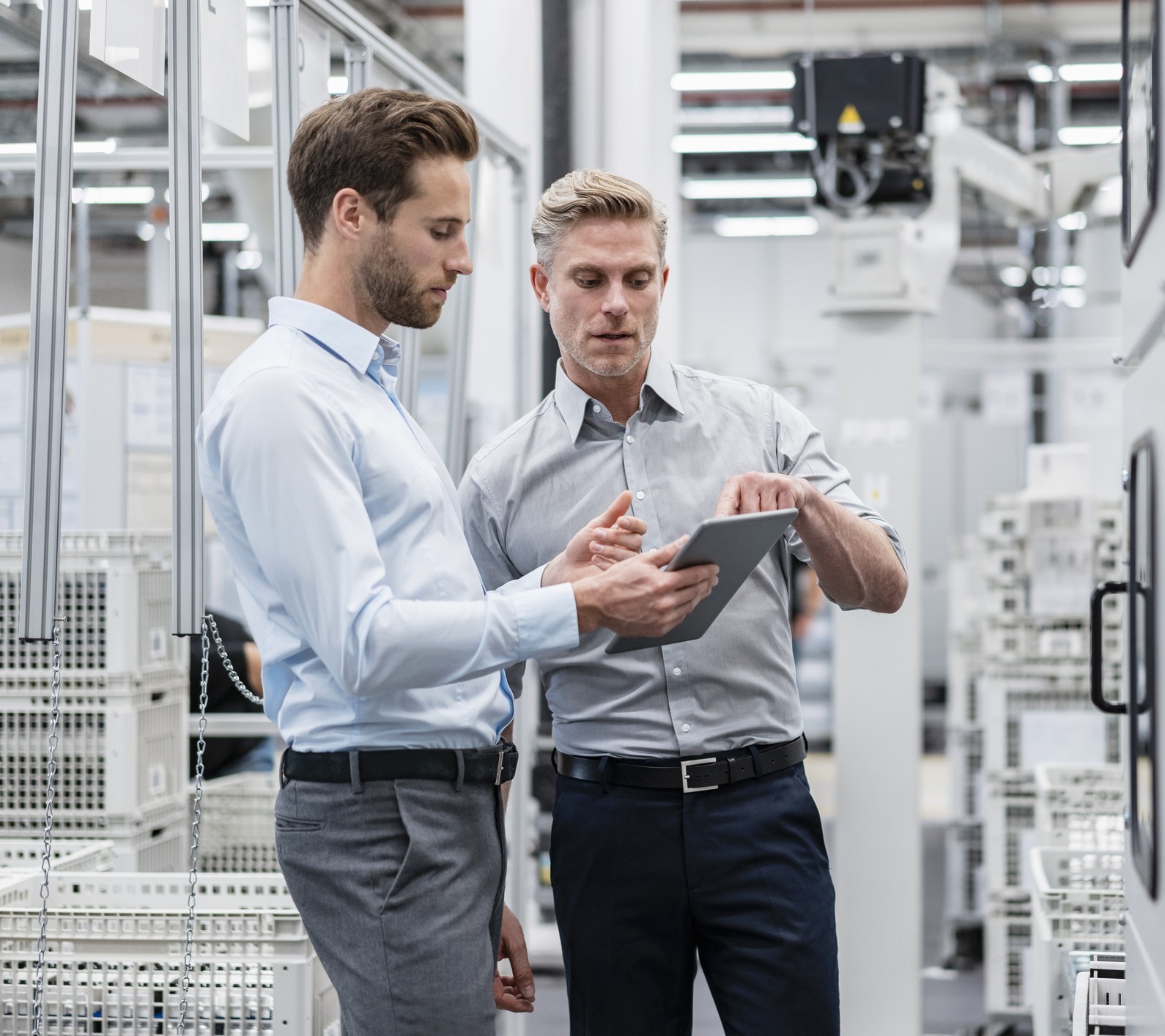Two men in business attire stand in a modern industrial facility, discussing information on a tablet. They are surrounded by white equipment and machinery, suggesting a high-tech manufacturing environment.
