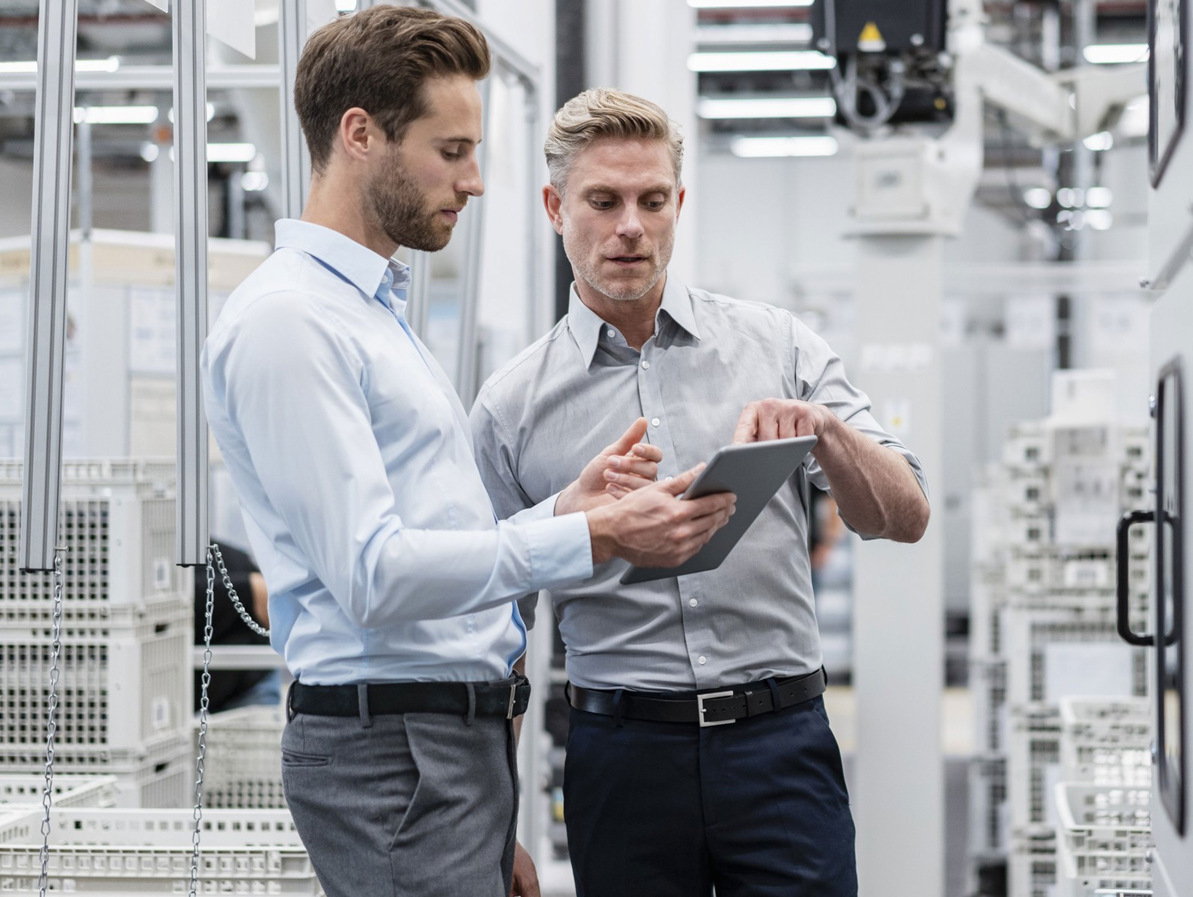 Two men in business attire stand in a modern industrial facility, discussing information on a tablet. They are surrounded by white equipment and machinery, suggesting a high-tech manufacturing environment.