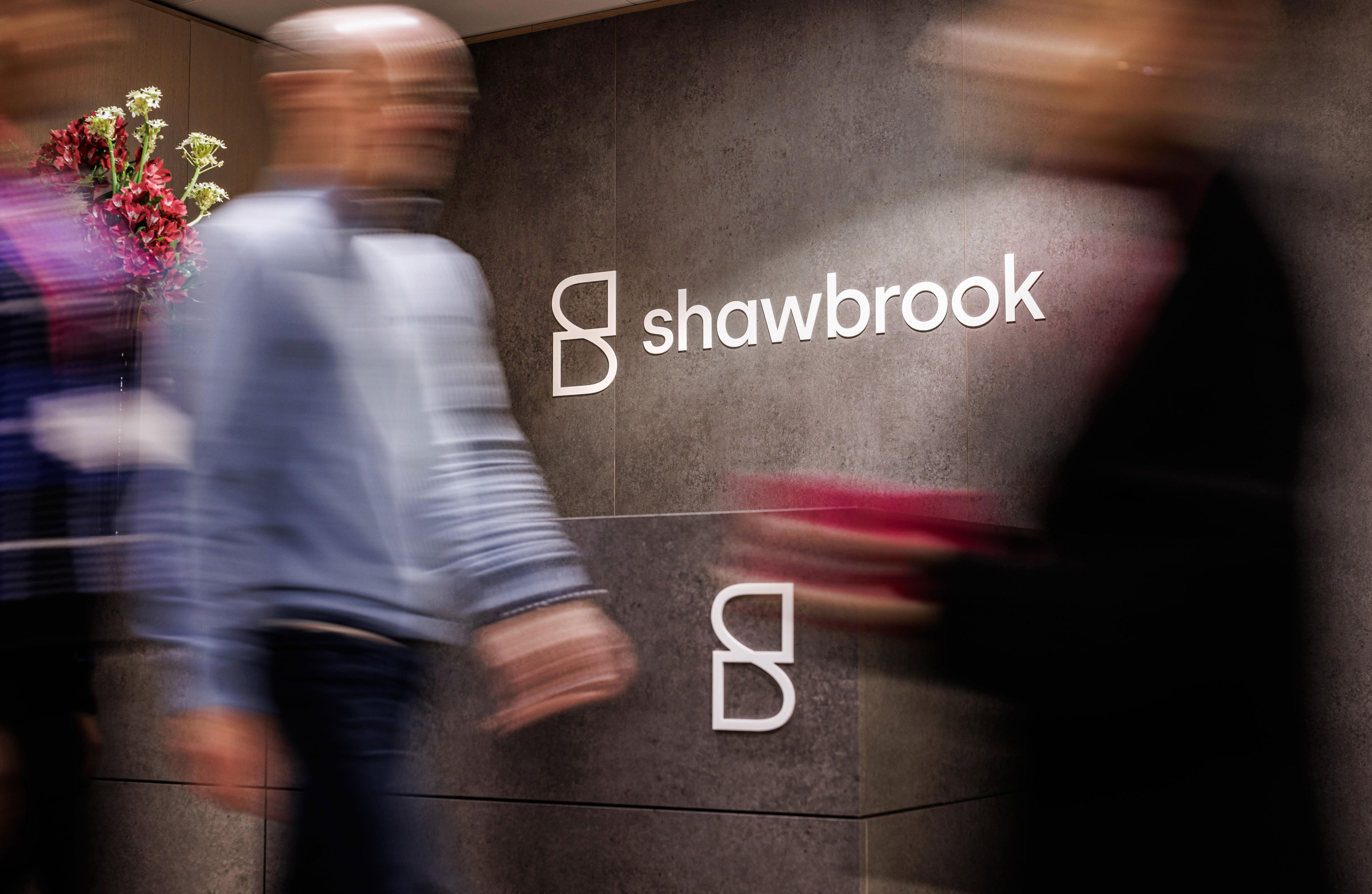 People walking past a wall with the "Shawbrook" logo on it. The scene is slightly blurry due to motion. A floral arrangement is visible in the background, adding a touch of color to the otherwise gray-toned environment.