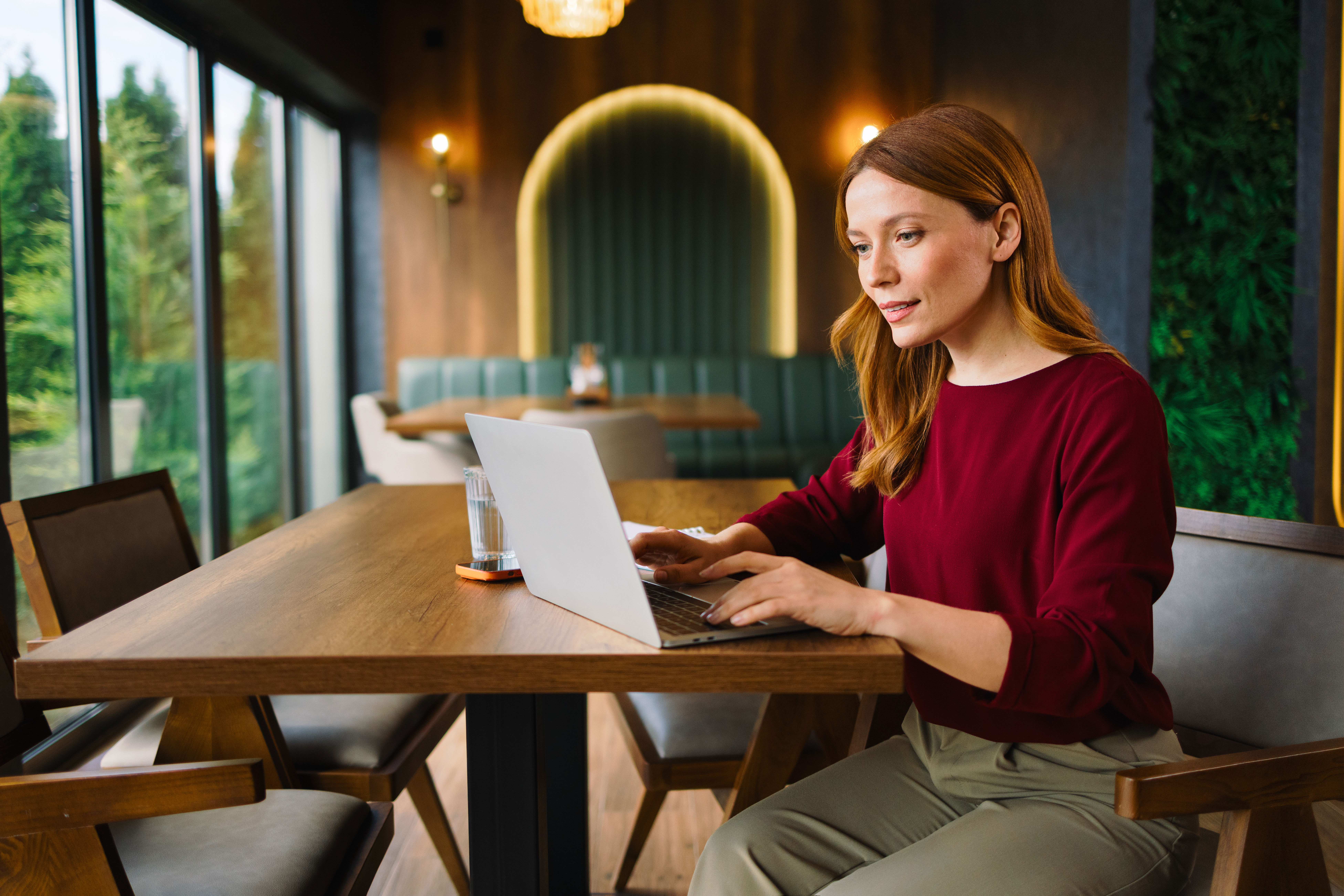 Woman with long red hair works on a laptop at a wooden table in a modern, well-lit cafe with large windows, green wall, and cozy booth seating in the background.