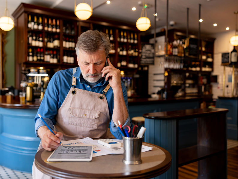 A man wearing an apron sits at a small table in a bar or restaurant, talking on the phone and reviewing paperwork, with shelves of bottles visible in the background.