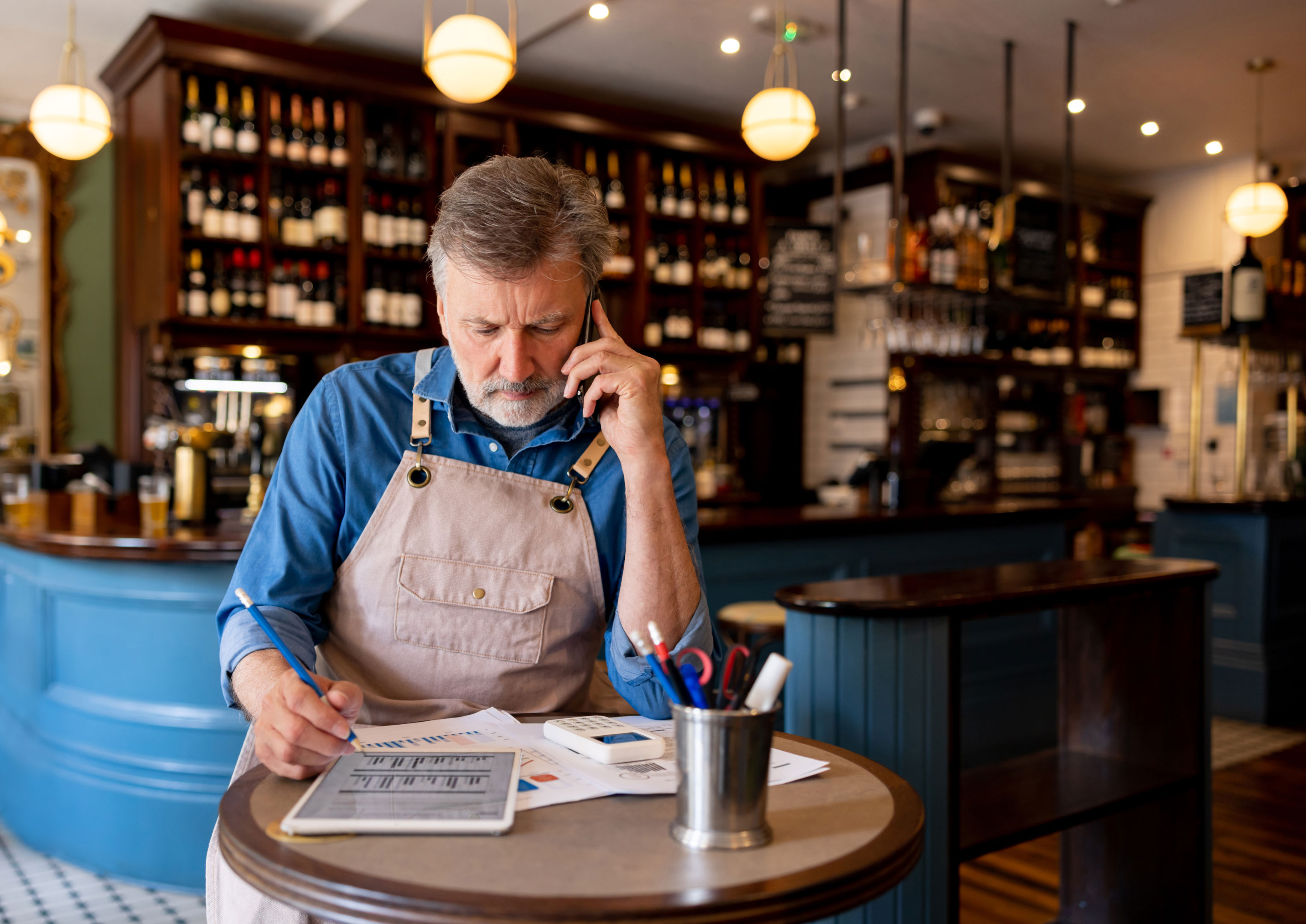A man wearing an apron sits at a small table in a bar or restaurant, talking on the phone and reviewing paperwork, with shelves of bottles visible in the background.
