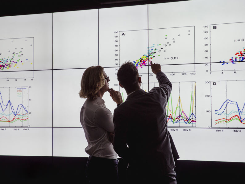 Two people stand in front of a large screen displaying colorful graphs and scatter plots, discussing the data and pointing at specific chart details.