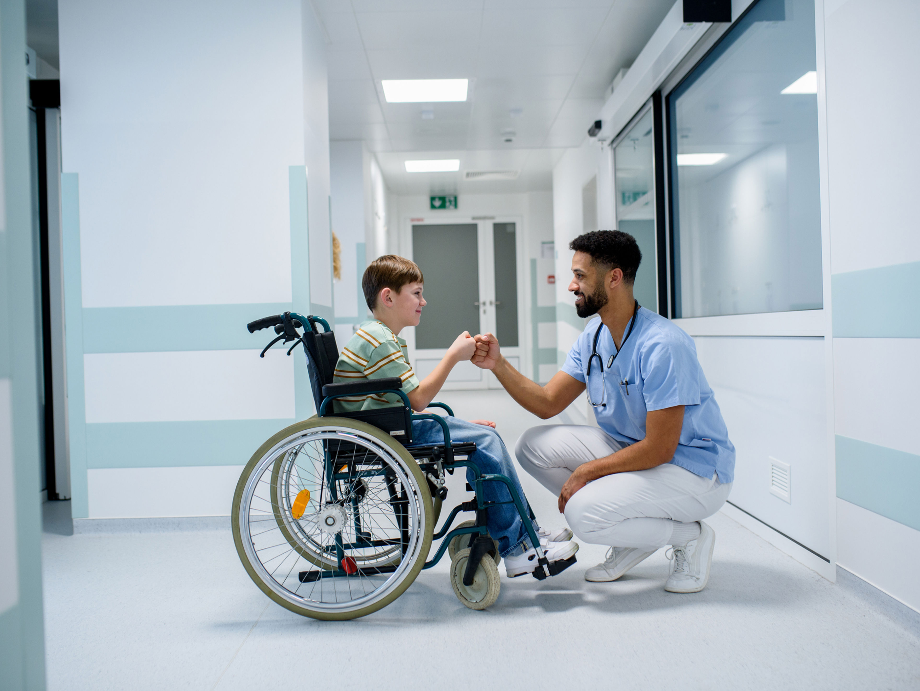 A young boy in a wheelchair and a male healthcare worker are smiling and greeting each other with a fist bump in a hospital corridor. The healthcare worker is squatting to be at the boy’s level.