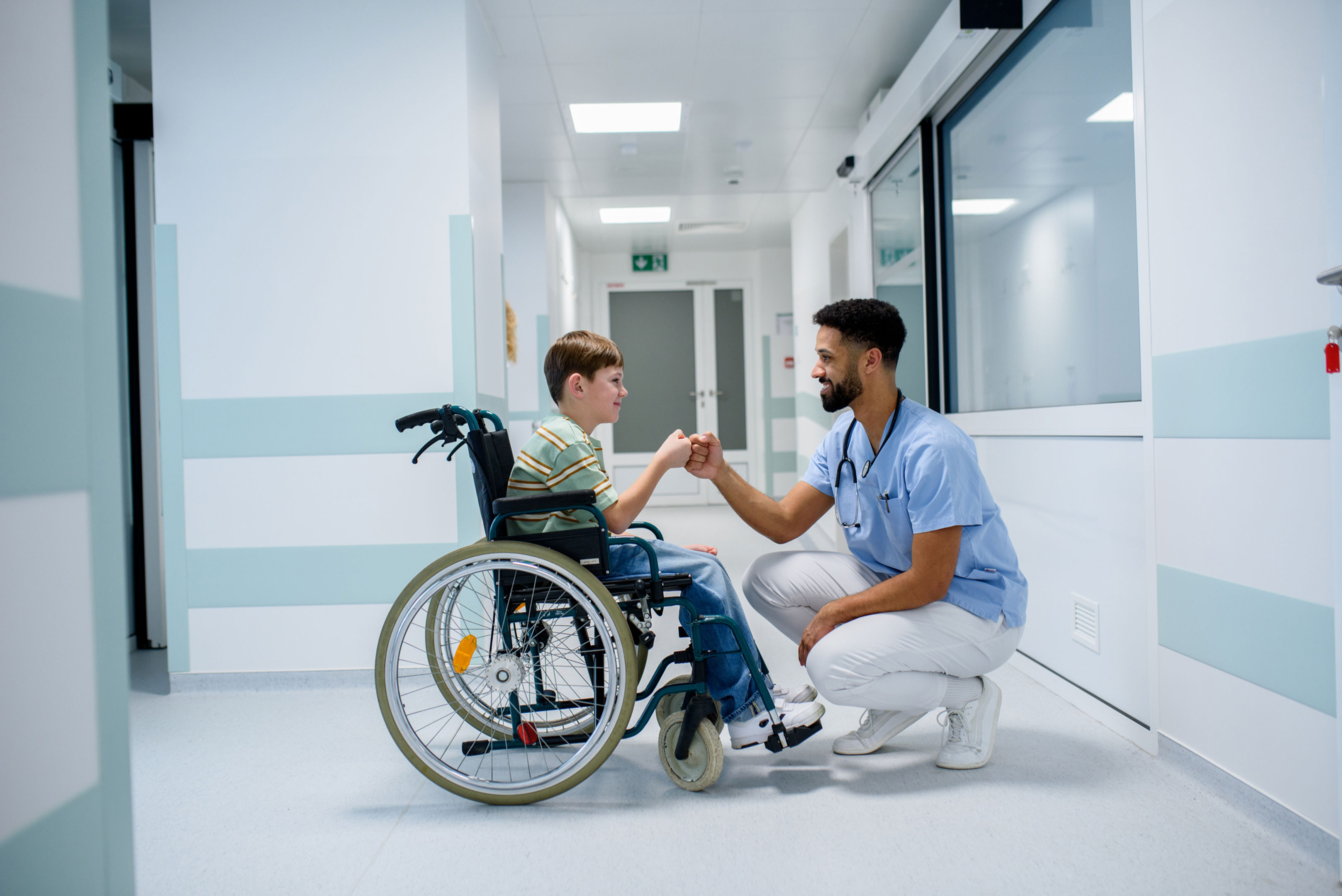 A young boy in a wheelchair and a male healthcare worker are smiling and greeting each other with a fist bump in a hospital corridor. The healthcare worker is squatting to be at the boy’s level.