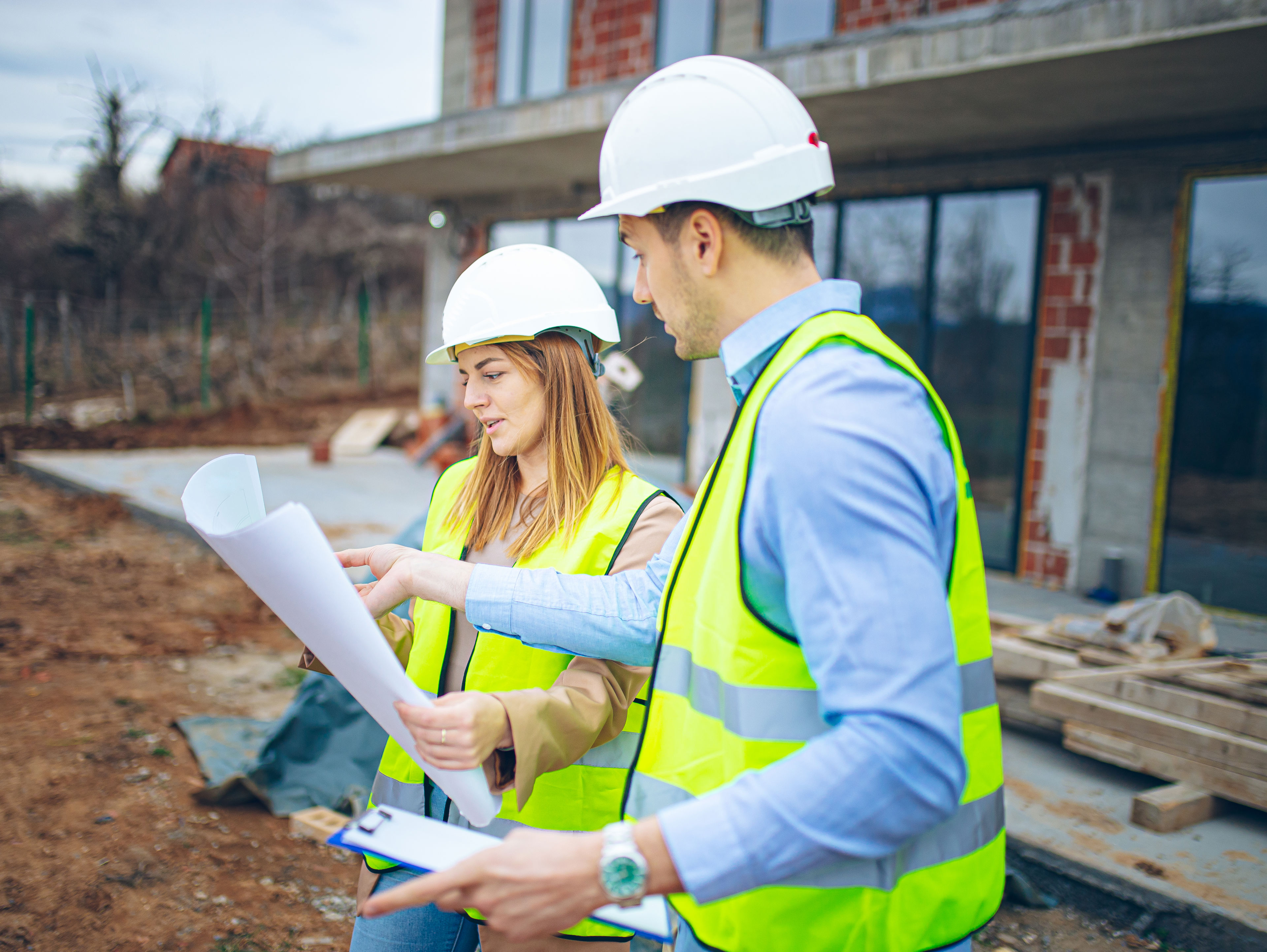 Two construction workers wearing hard hats and reflective vests review blueprints and documents at a building site, with a partially constructed structure visible in the background.