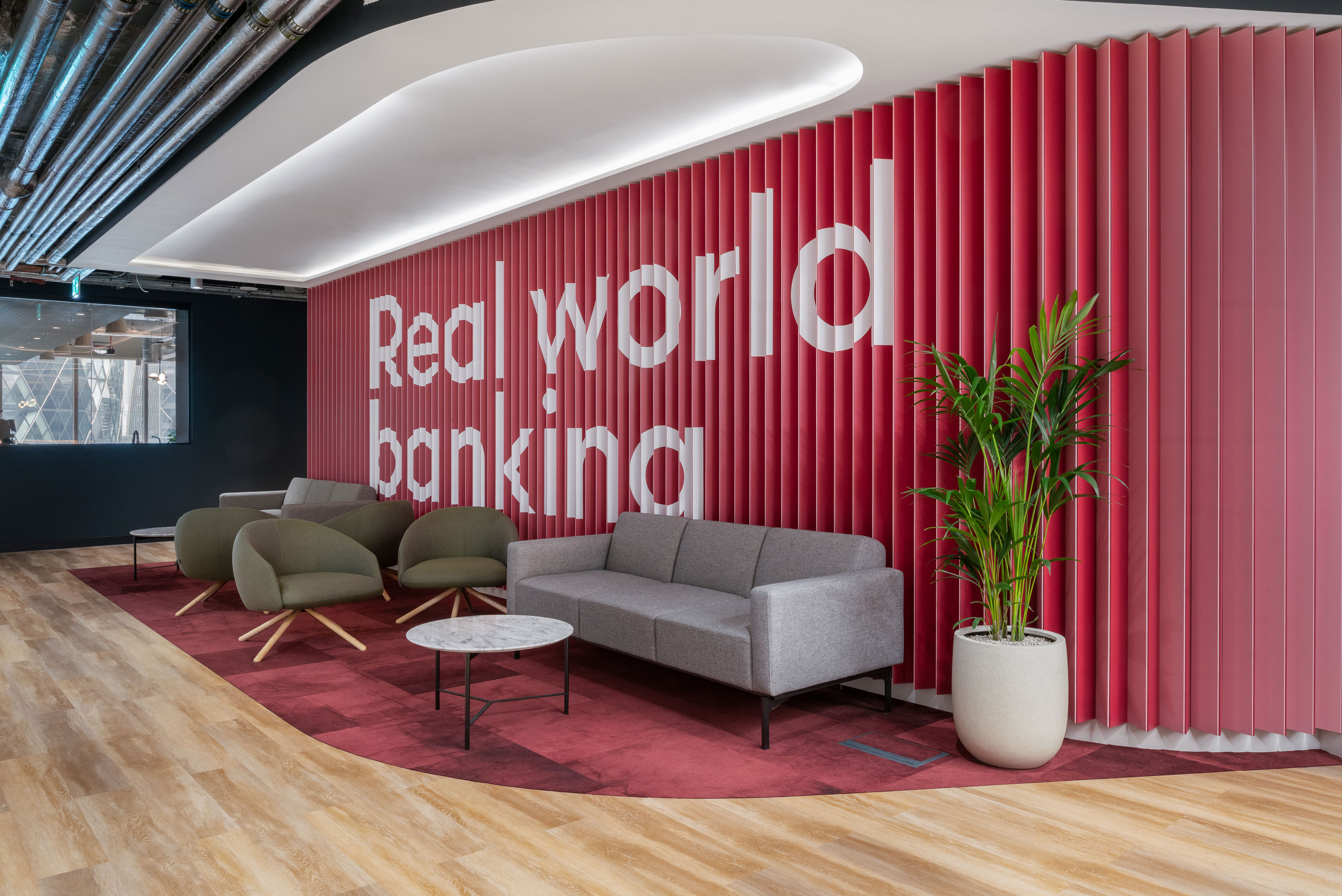 A modern office lounge with pink vertical wall panels displaying the words "Real world banking," gray and green chairs, a sofa, small tables, a potted plant, and wood flooring.