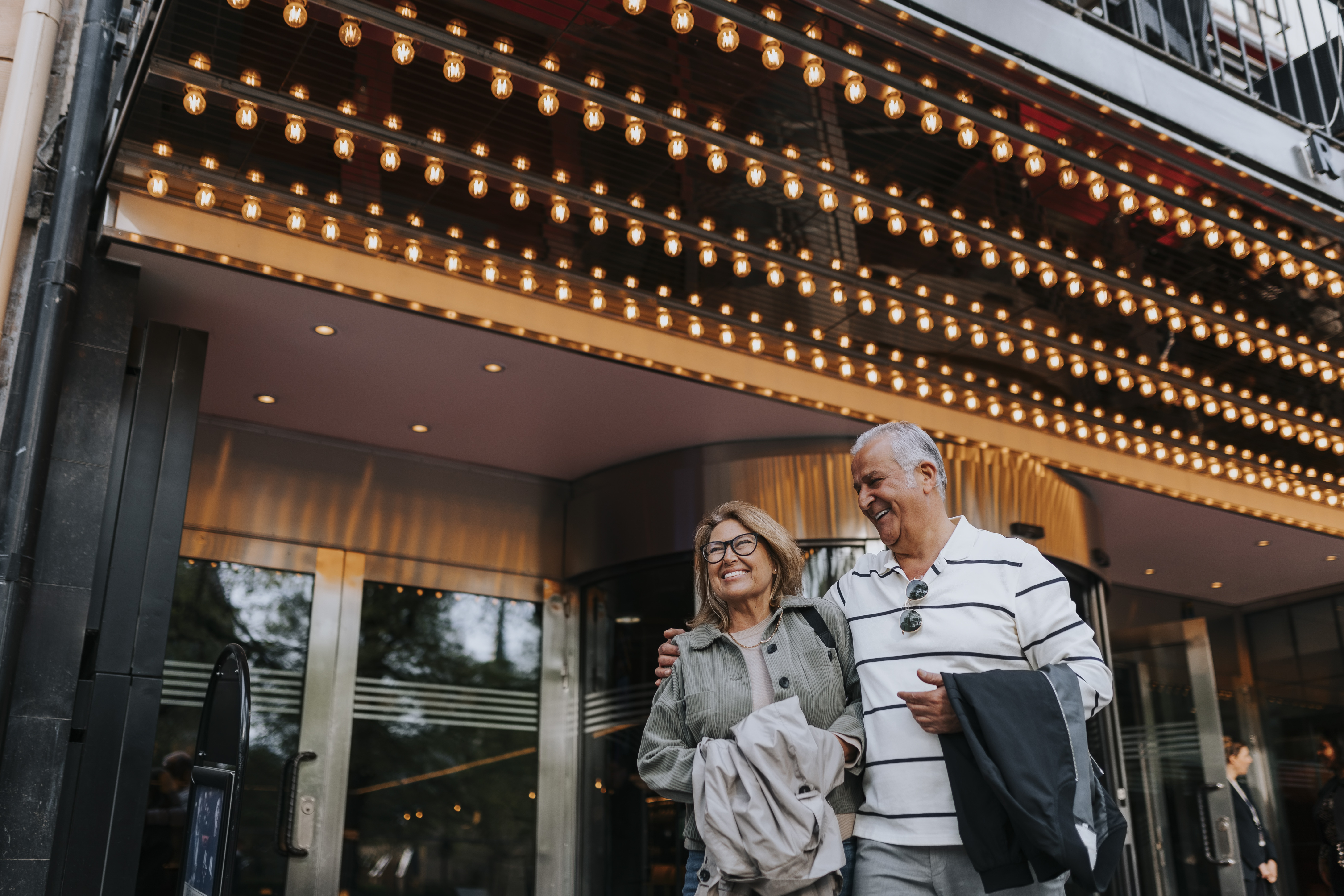 An older couple smiles and walks arm in arm under a brightly lit theater marquee, wearing casual clothes and holding jackets, with glass doors and reflections behind them.