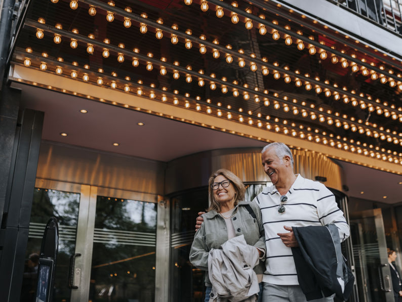 An older couple smiles and walks arm in arm under a brightly lit theater marquee, wearing casual clothes and holding jackets, with glass doors and reflections behind them.