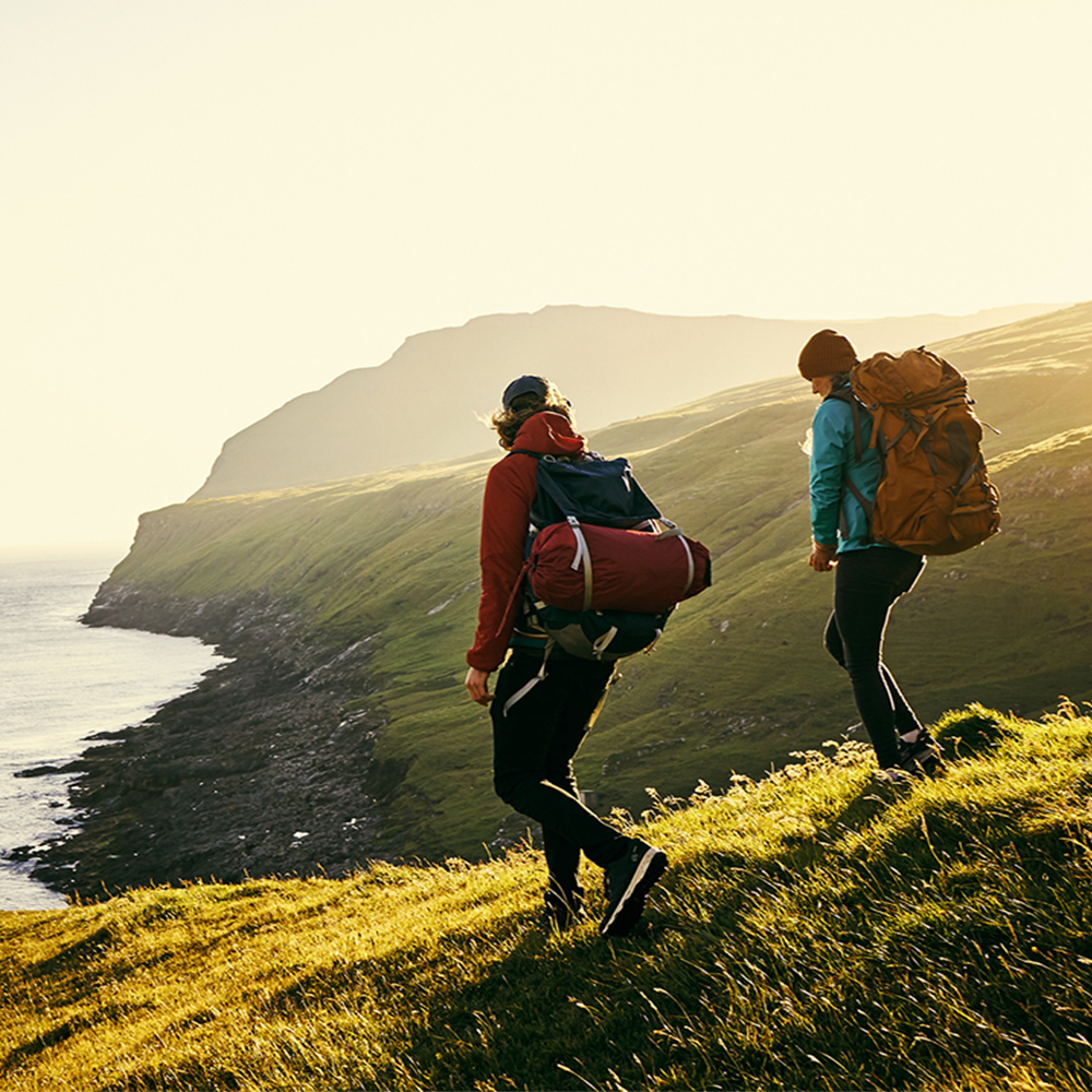 Two people with backpacks hike along a grassy coastal hillside at sunset, overlooking cliffs and the sea.
