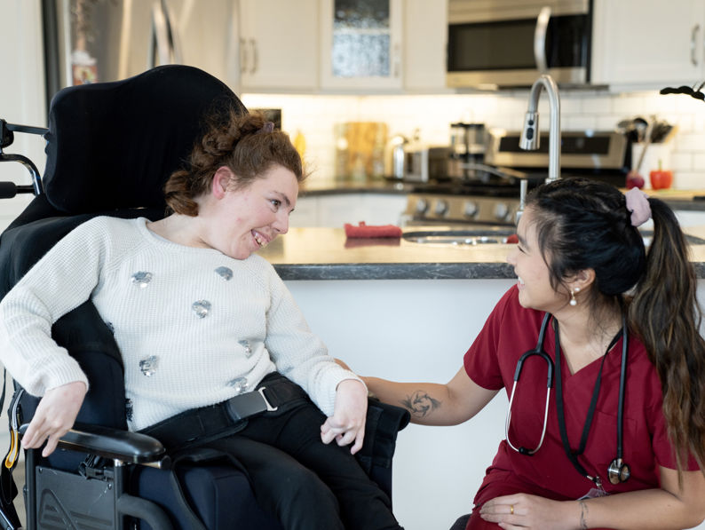 A woman in a wheelchair smiles and interacts warmly with a healthcare worker in red scrubs, who is kneeling beside her in a bright, modern kitchen.
