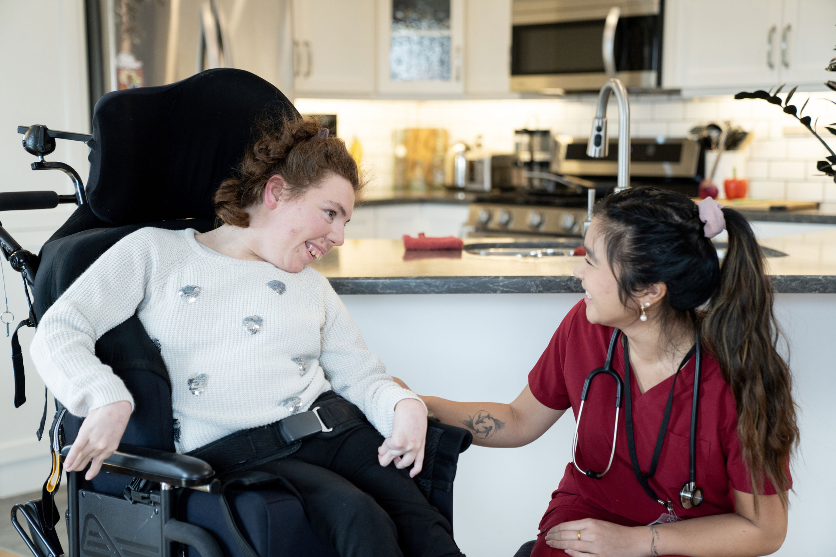 A woman in a wheelchair smiles and interacts warmly with a healthcare worker in red scrubs, who is kneeling beside her in a bright, modern kitchen.