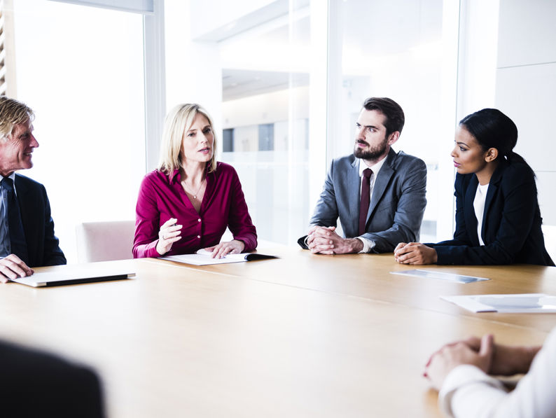 Five professionals sit around a conference table in a bright office, engaged in discussion. One woman in a red blouse is speaking while others, dressed in business attire, listen attentively.