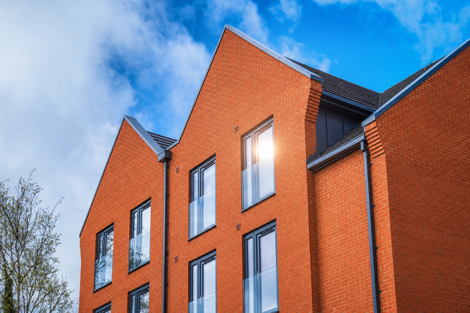 Modern red brick building with three peaked roofs, large rectangular windows, and glass balconies, set against a blue sky with scattered clouds. A tree is partially visible on the left side.