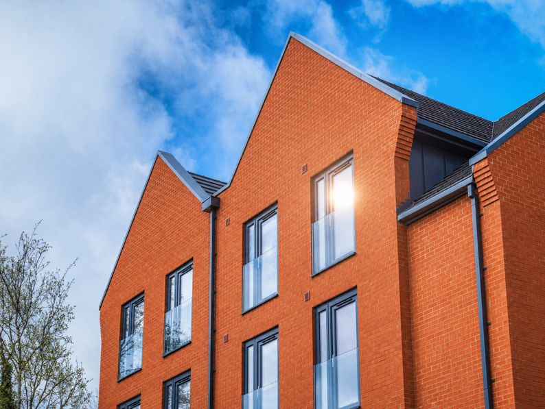 Modern red brick building with three peaked roofs, large rectangular windows, and glass balconies, set against a blue sky with scattered clouds. A tree is partially visible on the left side.