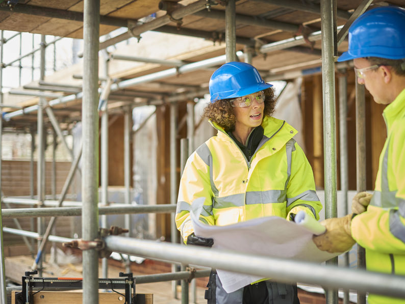 Two construction workers in high-visibility jackets and blue hard hats discuss plans at a building site surrounded by scaffolding. One holds a blueprint while the other listens attentively.
