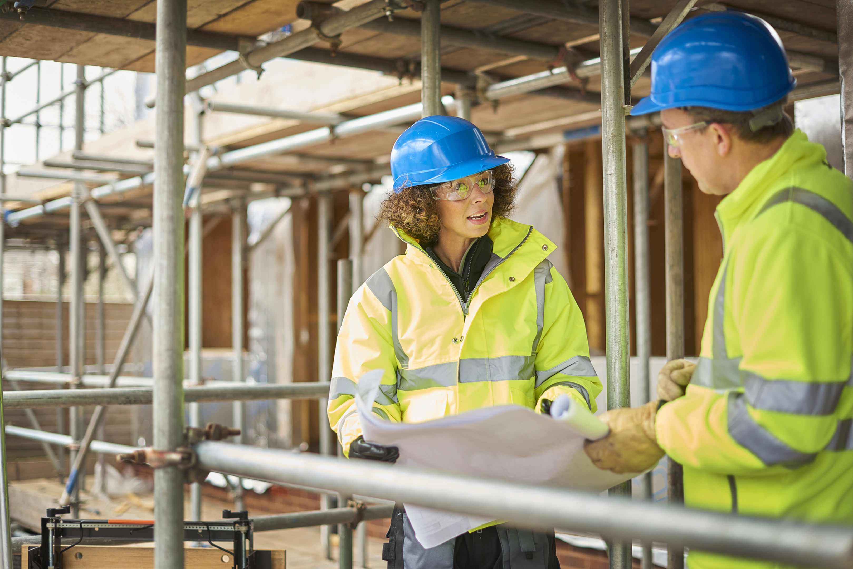Two construction workers in high-visibility jackets and blue hard hats discuss plans at a building site surrounded by scaffolding. One holds a blueprint while the other listens attentively.