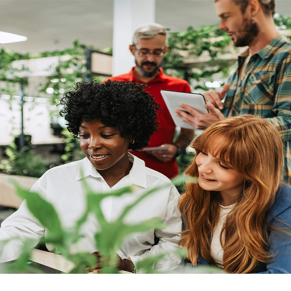 Four adults collaborate in a bright, modern office with green plants. Two women sit at a table smiling, while two men stand behind them, one holding a tablet and the other writing on a notepad.