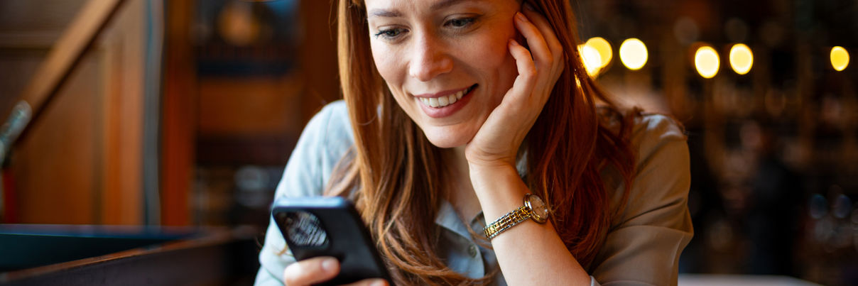 A woman with reddish-brown hair smiles while looking at her smartphone in a cozy cafe. She wears a light blouse and a watch, with a coffee cup on the table in front of her. Warm lights are blurred in the background.
