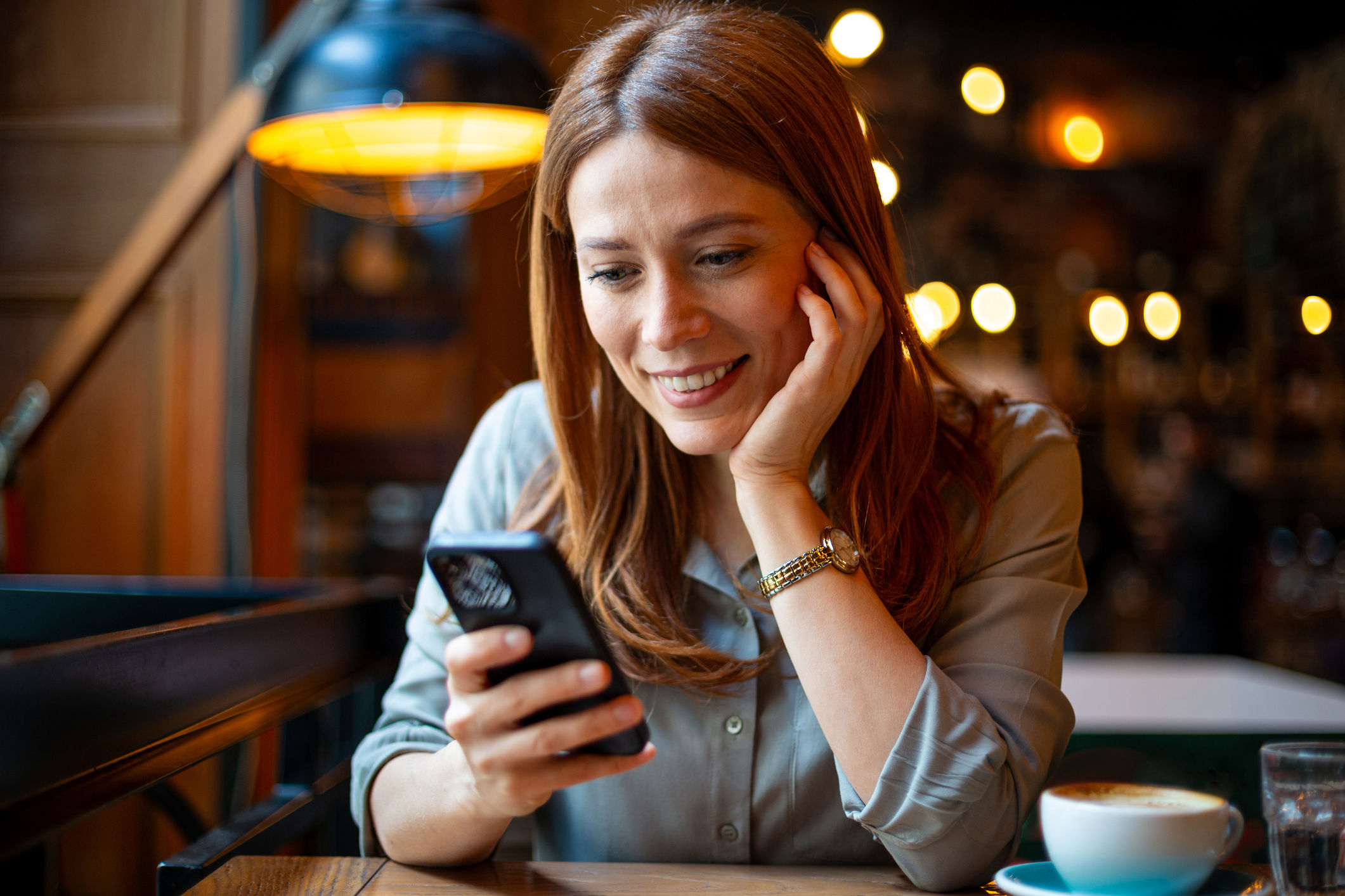 A woman with reddish-brown hair smiles while looking at her smartphone in a cozy cafe. She wears a light blouse and a watch, with a coffee cup on the table in front of her. Warm lights are blurred in the background.