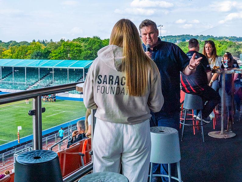 A group of people socialize on an outdoor balcony overlooking a rugby stadium. One person wears a hoodie labeled "Saracens Foundation." The pitch and empty stands are visible in the background under a partly cloudy sky.