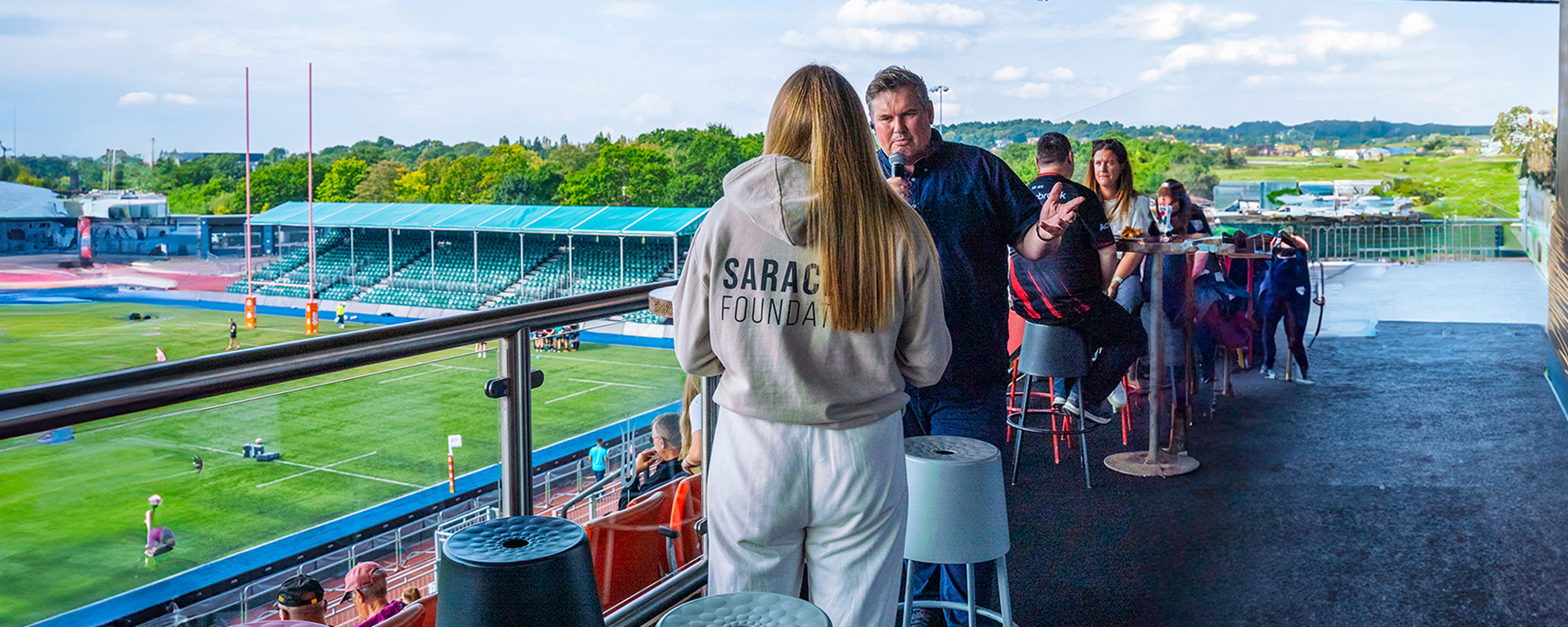 A group of people socialize on an outdoor balcony overlooking a rugby stadium. One person wears a hoodie labeled "Saracens Foundation." The pitch and empty stands are visible in the background under a partly cloudy sky.