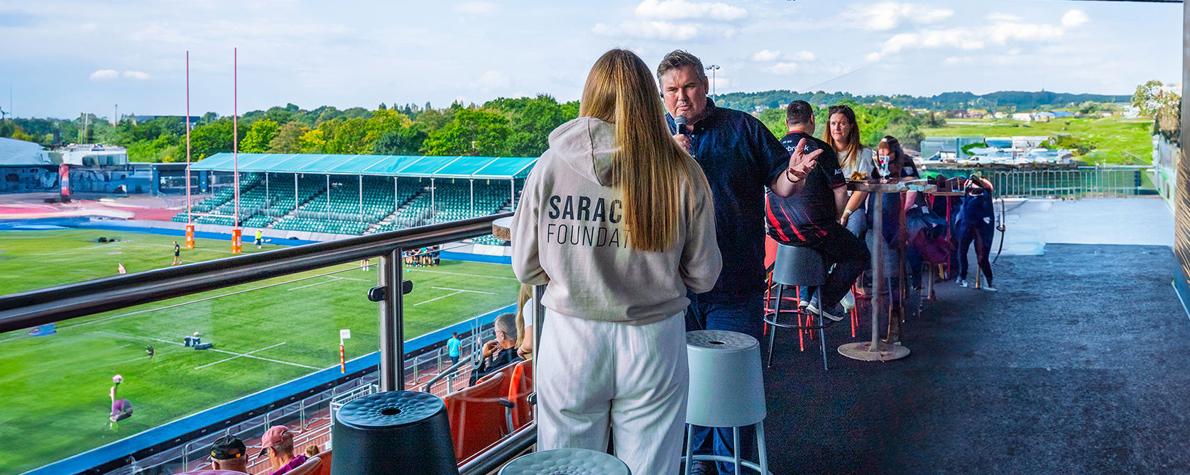 A group of people socialize on an outdoor balcony overlooking a rugby stadium. One person wears a hoodie labeled "Saracens Foundation." The pitch and empty stands are visible in the background under a partly cloudy sky.