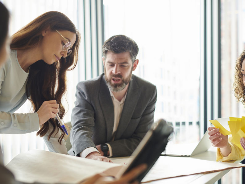 Four people sit and stand around a table in a well-lit office, engaged in discussion. One person points at a large sheet of paper, while another holds several yellow sticky notes.