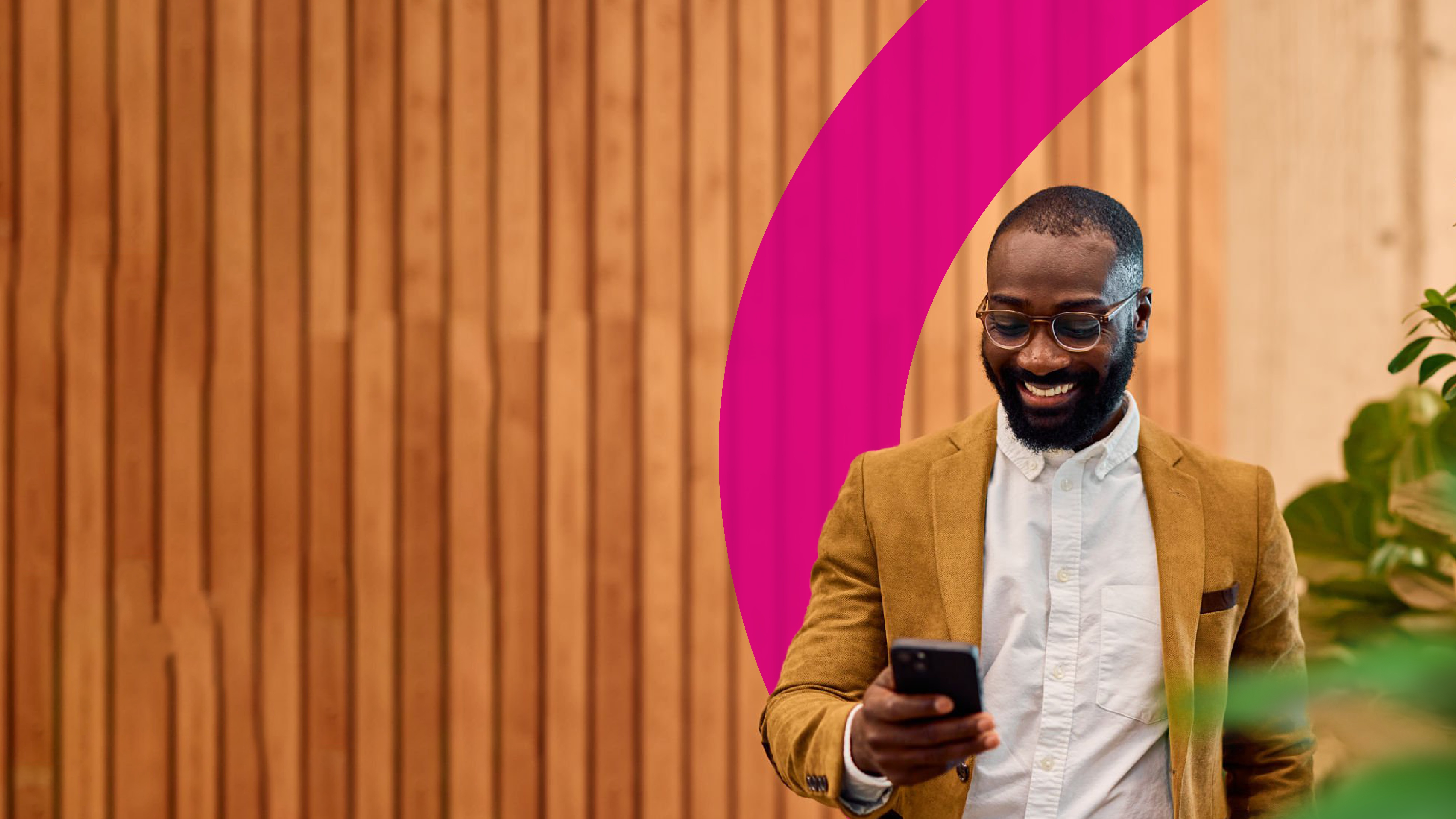 A smiling man in a tan blazer and glasses looks at his phone while standing in front of a wooden wall with a pink curved design and some green plants nearby.
