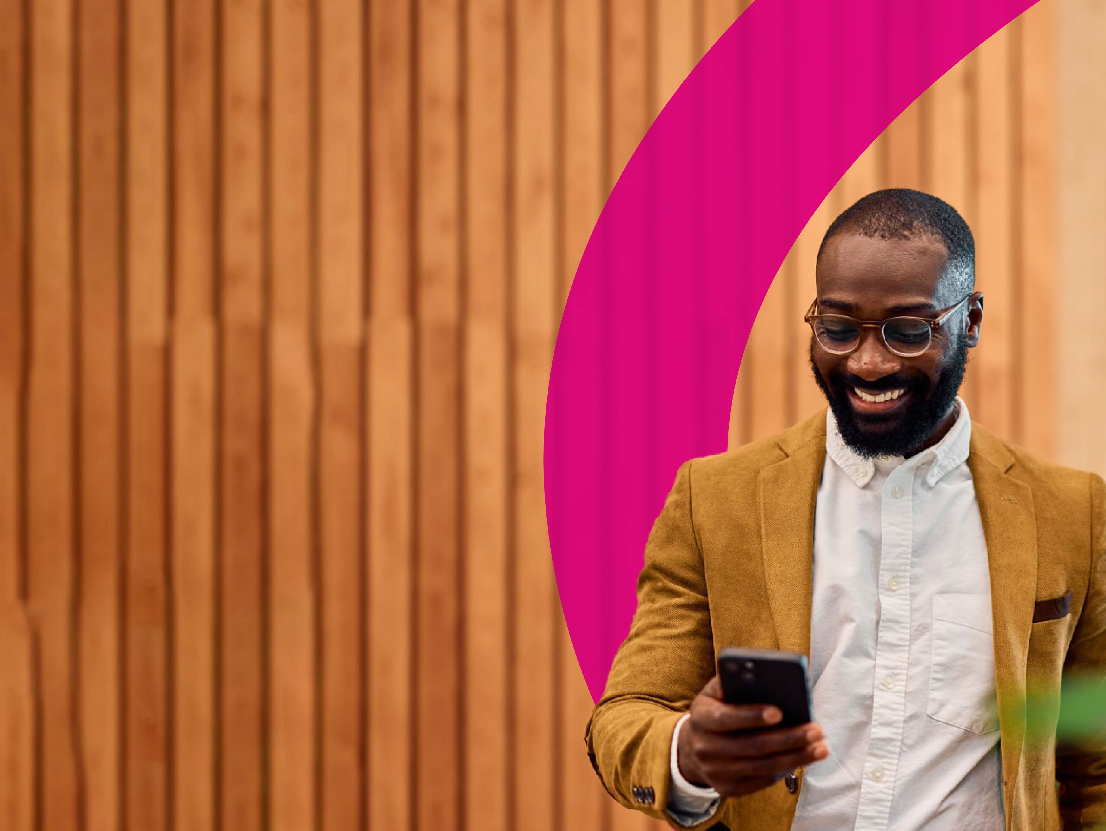A smiling man in a tan blazer and glasses looks at his phone while standing in front of a wooden wall with a pink curved design and some green plants nearby.