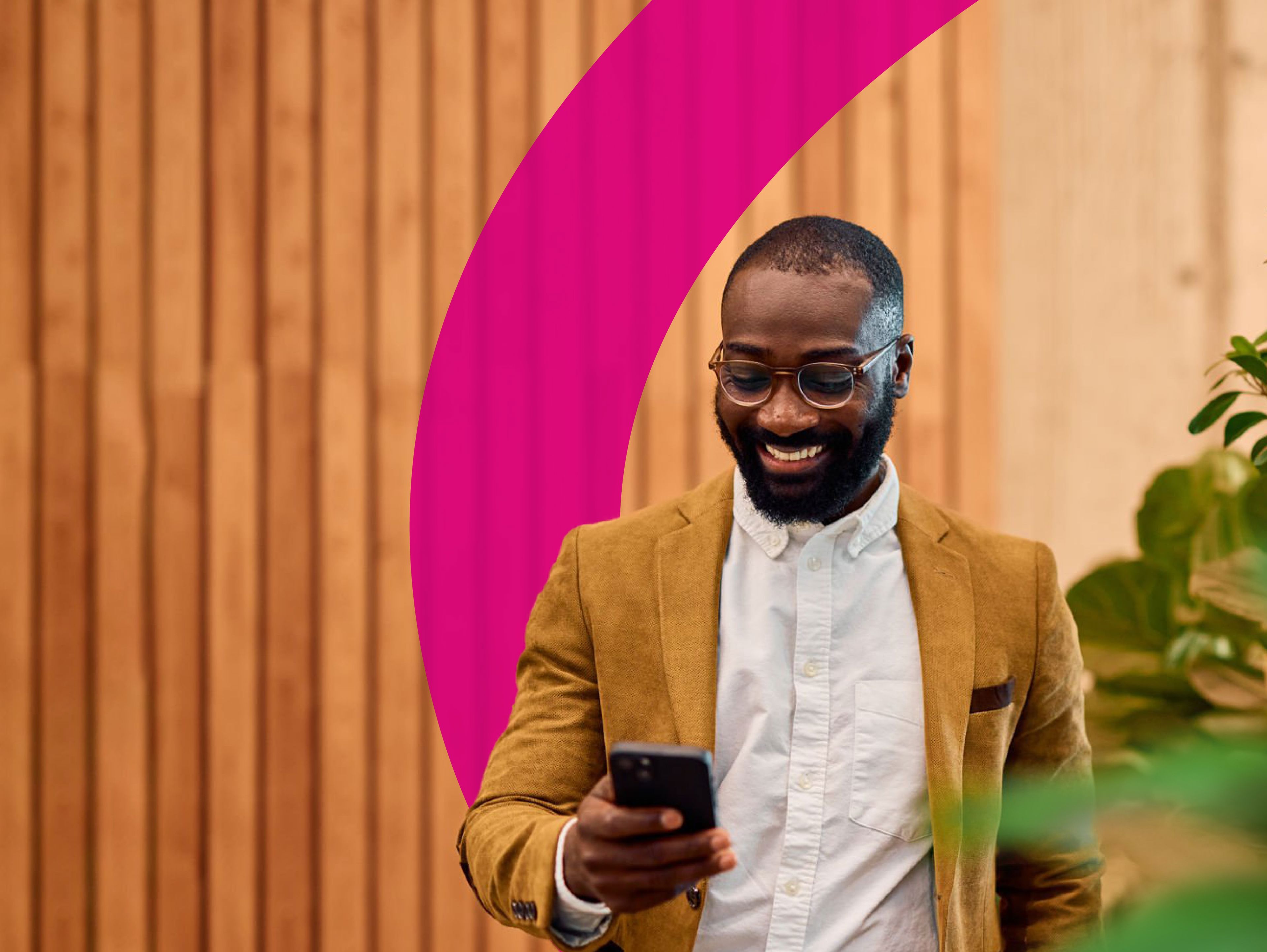 A smiling man in a tan blazer and glasses looks at his phone while standing in front of a wooden wall with a pink curved design and some green plants nearby.