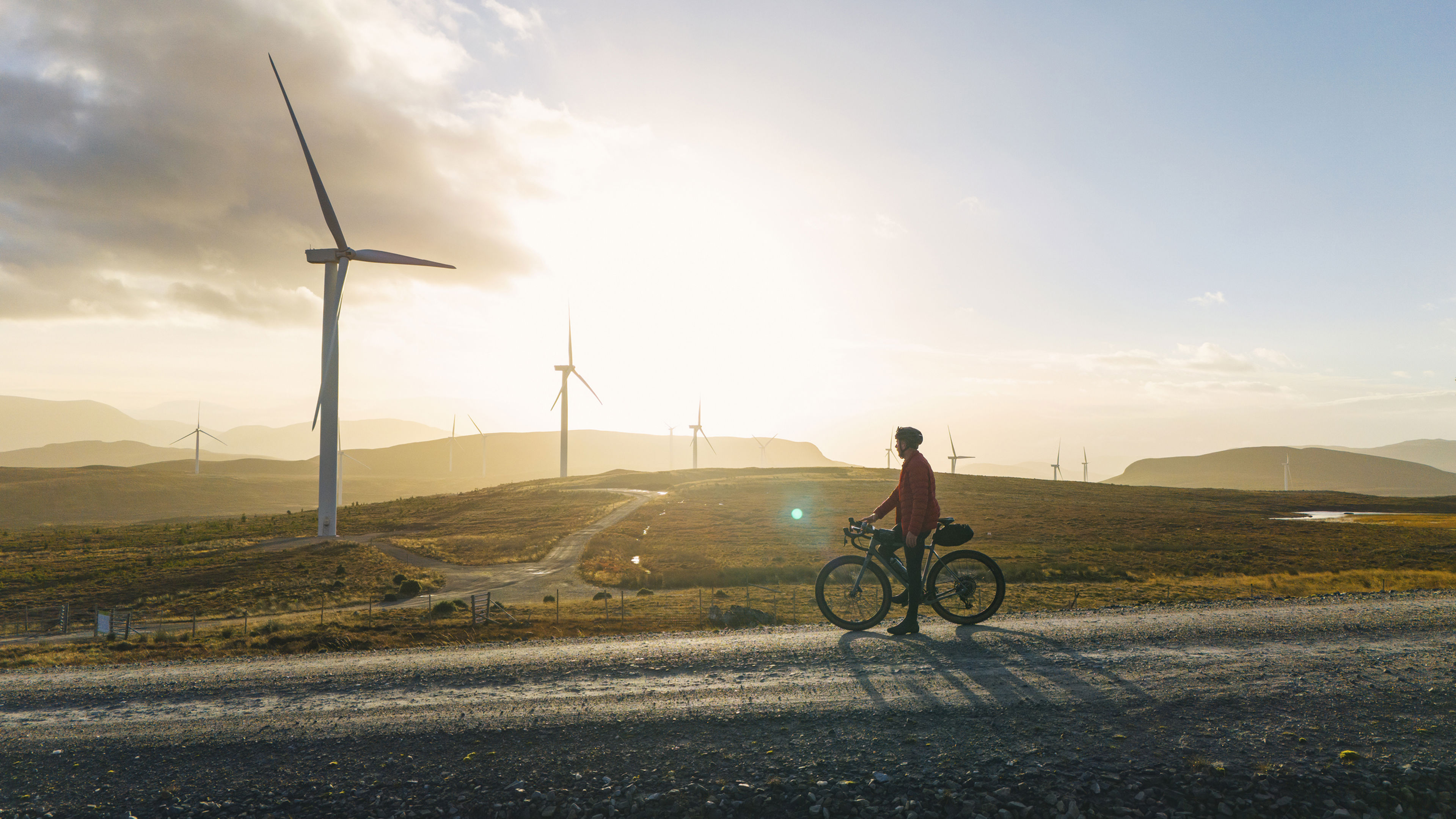 A person rides a bicycle on a gravel road through a landscape dotted with wind turbines during sunset, with golden sunlight illuminating the hills and sky in the background.