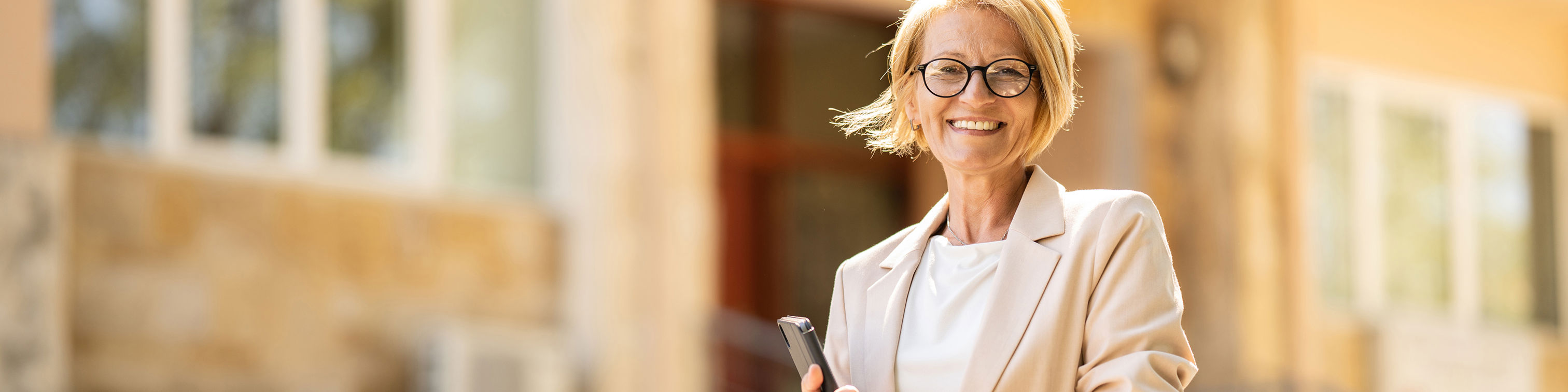 Smiling woman with short blonde hair and glasses, wearing a beige blazer, stands outside a building holding a tablet or notebook. The background features stairs, windows, and beige walls.