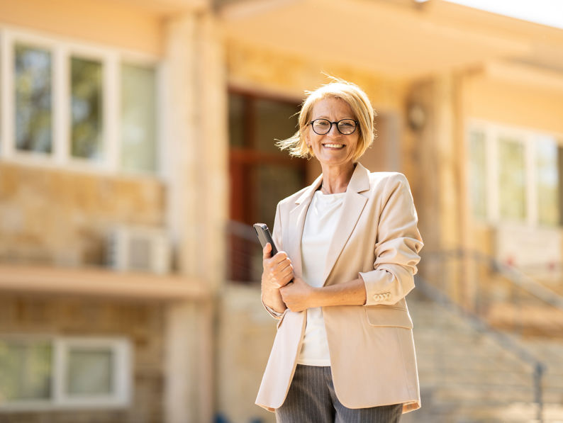 Smiling woman with short blonde hair and glasses, wearing a beige blazer, stands outside a building holding a tablet or notebook. The background features stairs, windows, and beige walls.