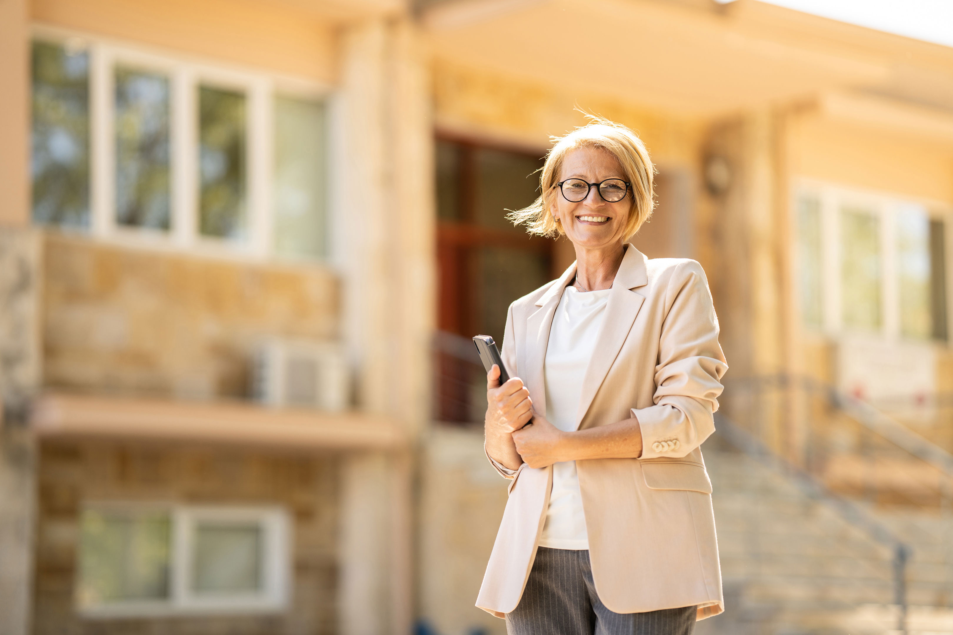 Smiling woman with short blonde hair and glasses, wearing a beige blazer, stands outside a building holding a tablet or notebook. The background features stairs, windows, and beige walls.