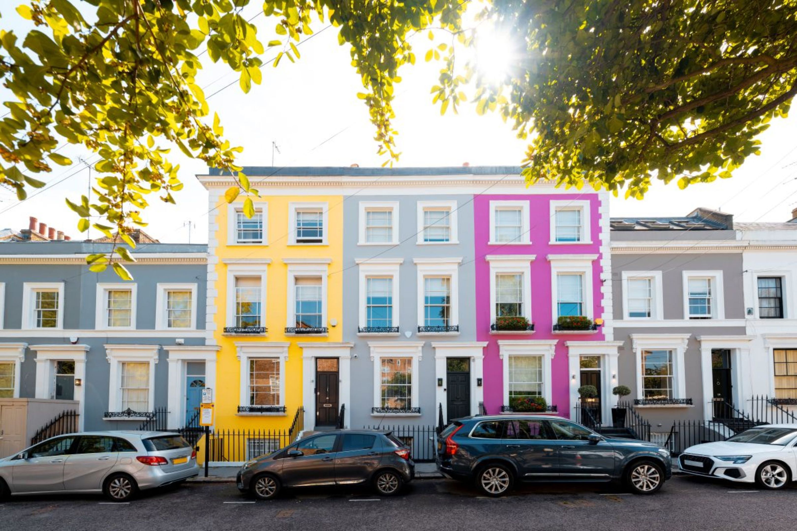 A row of colorful terraced houses, including bright yellow and pink facades, with several parked cars on the street and sunlight filtering through leafy tree branches above.