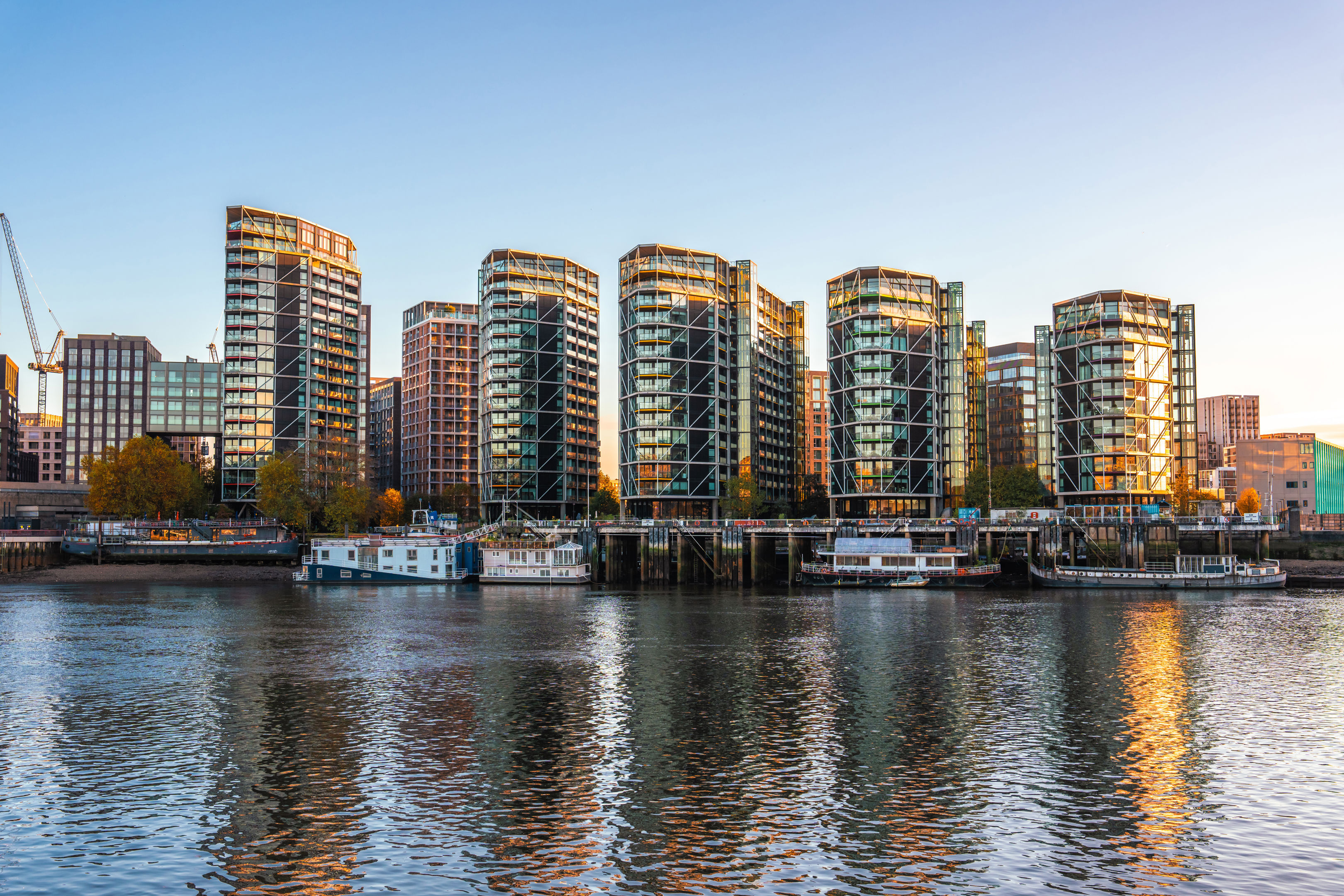 Modern glass apartment buildings line a waterfront, reflecting in the calm river below. Several houseboats are moored along the shore, and the sky is clear with warm light from the setting sun.