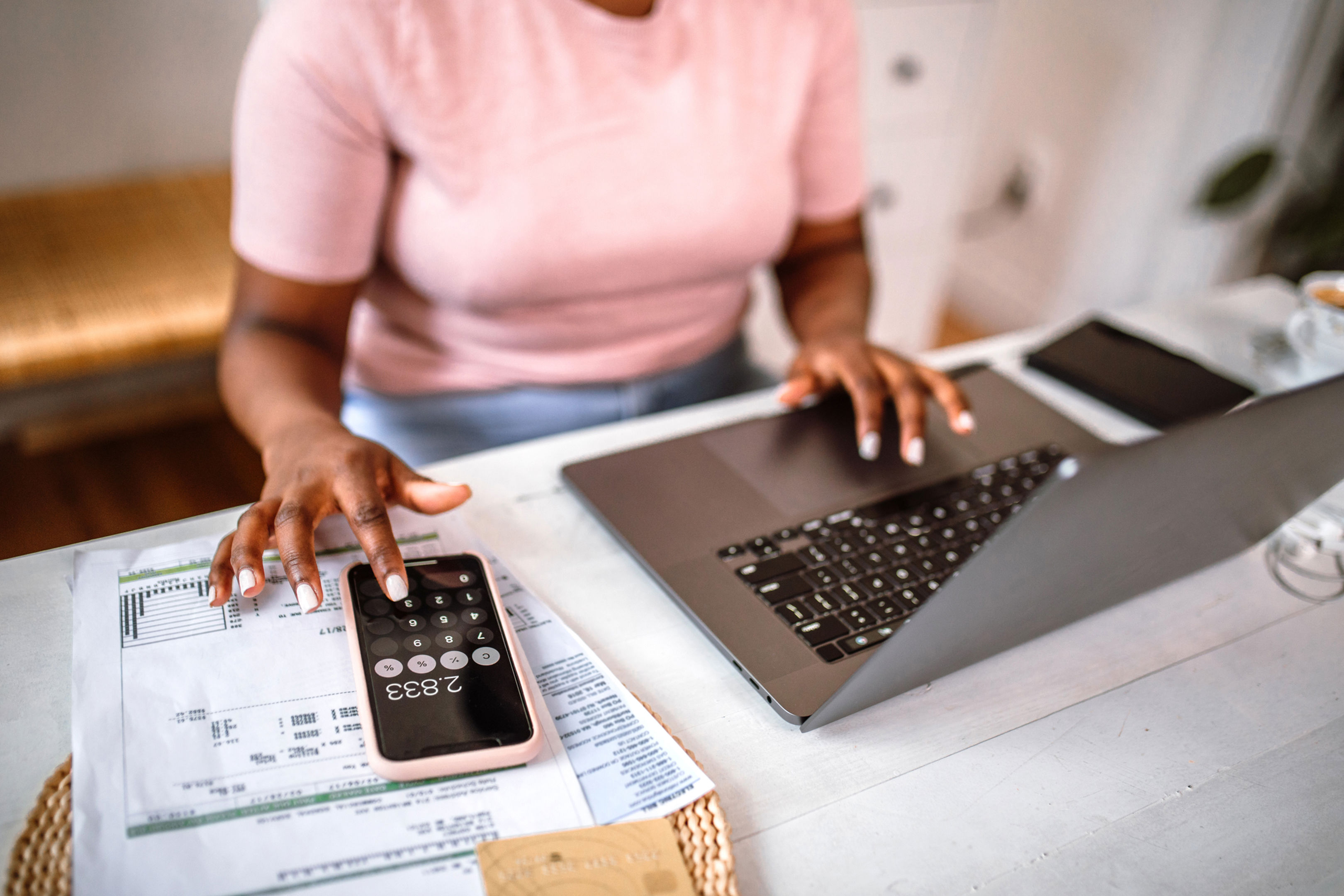 Women in pink top sitting at desk working on laptop of using calculator on her phone